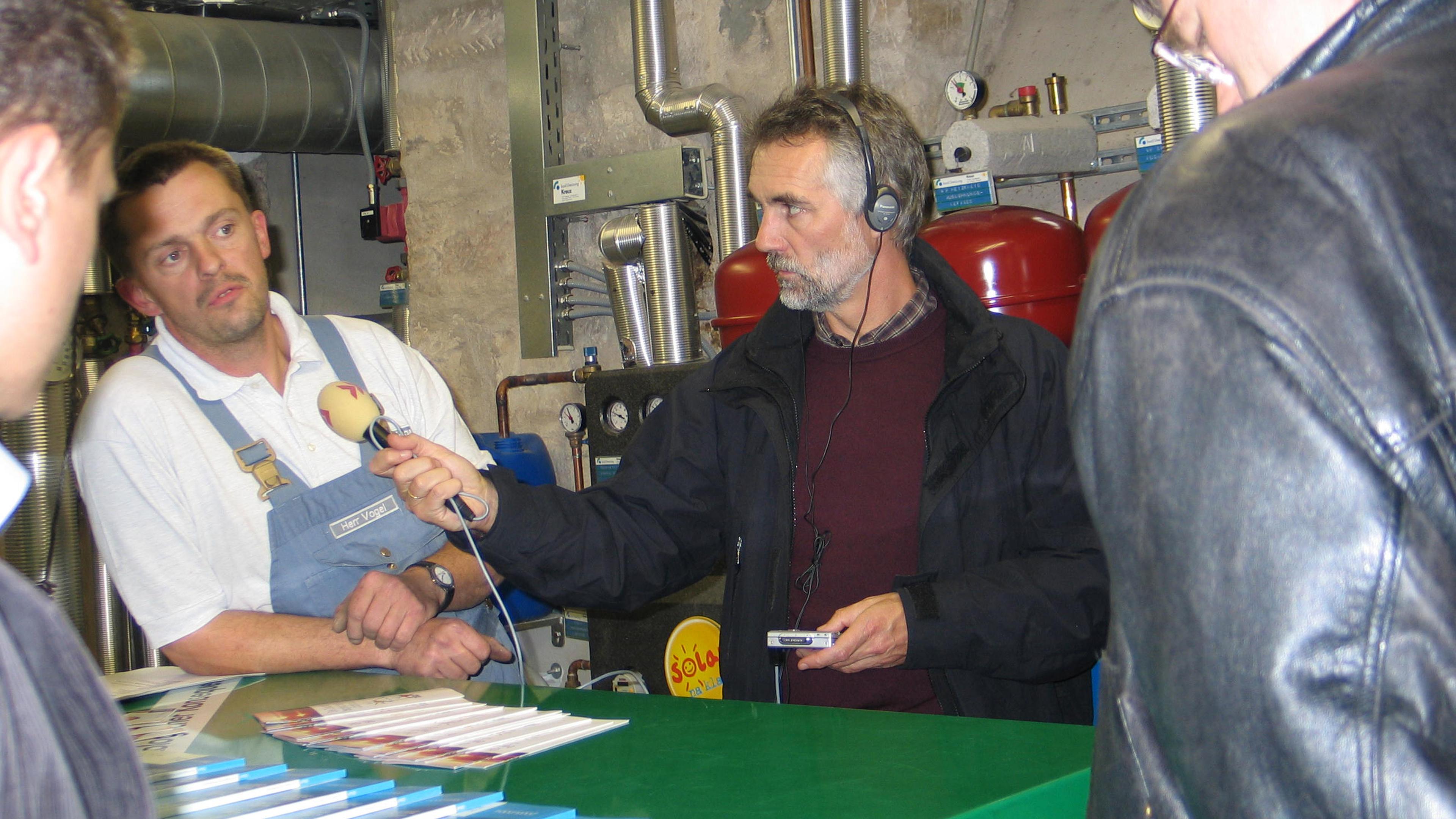 Ein Mann mit Kopfhörern interviewt einen anderen Mann in Arbeitskleidung mit einem Mikrofon, umgeben von Industrieanlagen und Broschüren auf einem grünen Tisch.