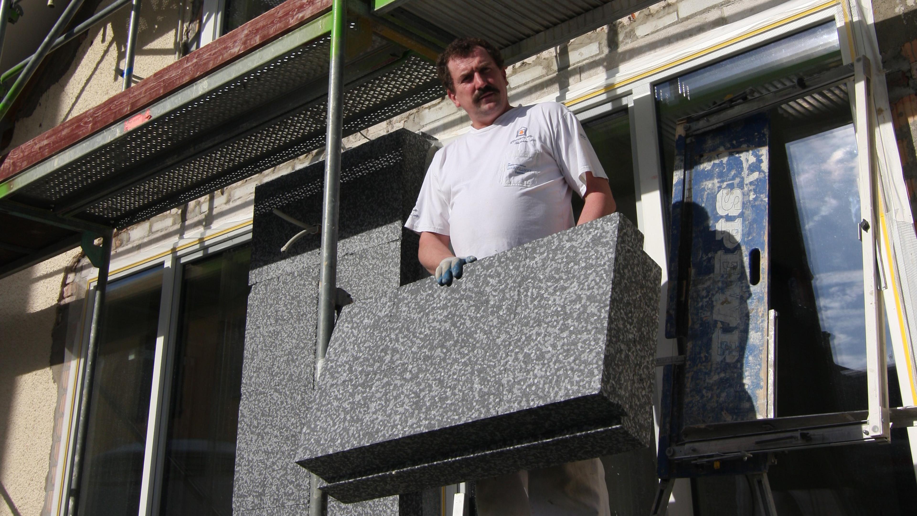Ein Mann in einem weißen Hemd, der auf einer Baustelle eine große graue Marmorplatte hält, im Hintergrund sind Gerüste und Baumaterialien zu sehen.