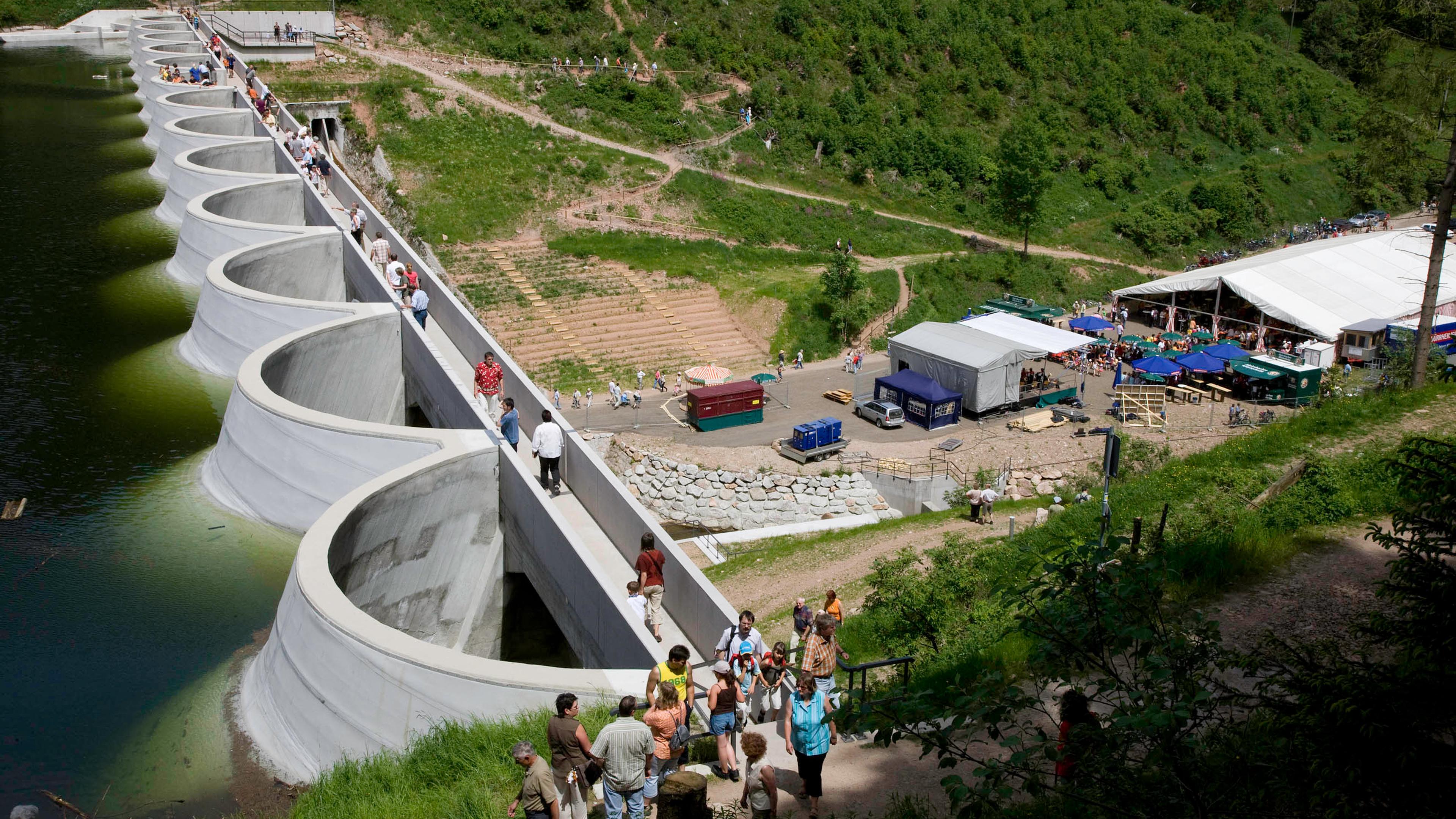 Menschen, die auf einer geschwungenen Dammbrücke neben einem Teich spazieren gehen, im Hintergrund Zelte und ein Versammlungsplatz auf einem grasbewachsenen Hügel.