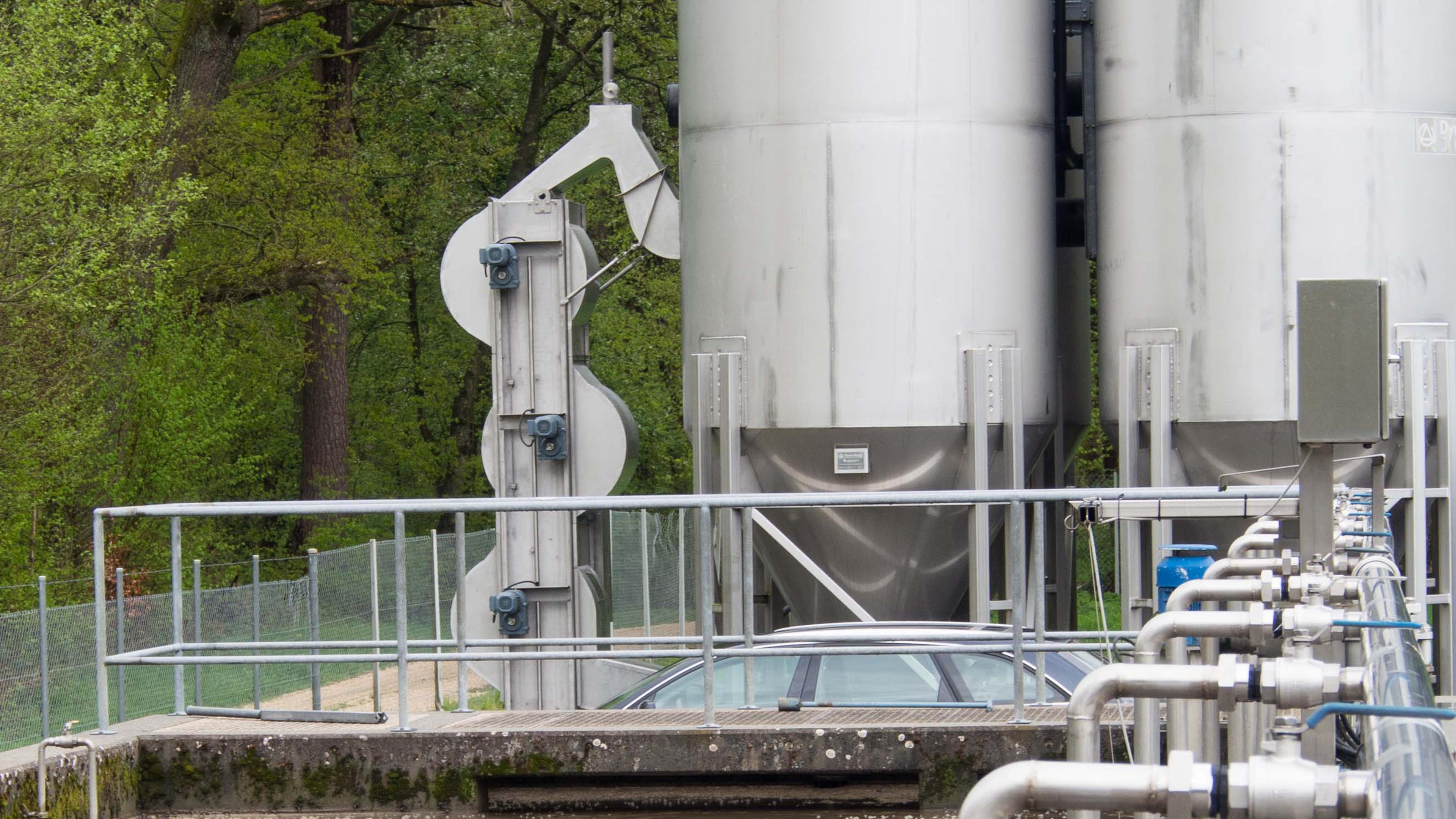 Industrietanks und Metallrohre in einem umzäunten Bereich am Waldrand. Hinter der Anlage ist teilweise ein Auto zu sehen.