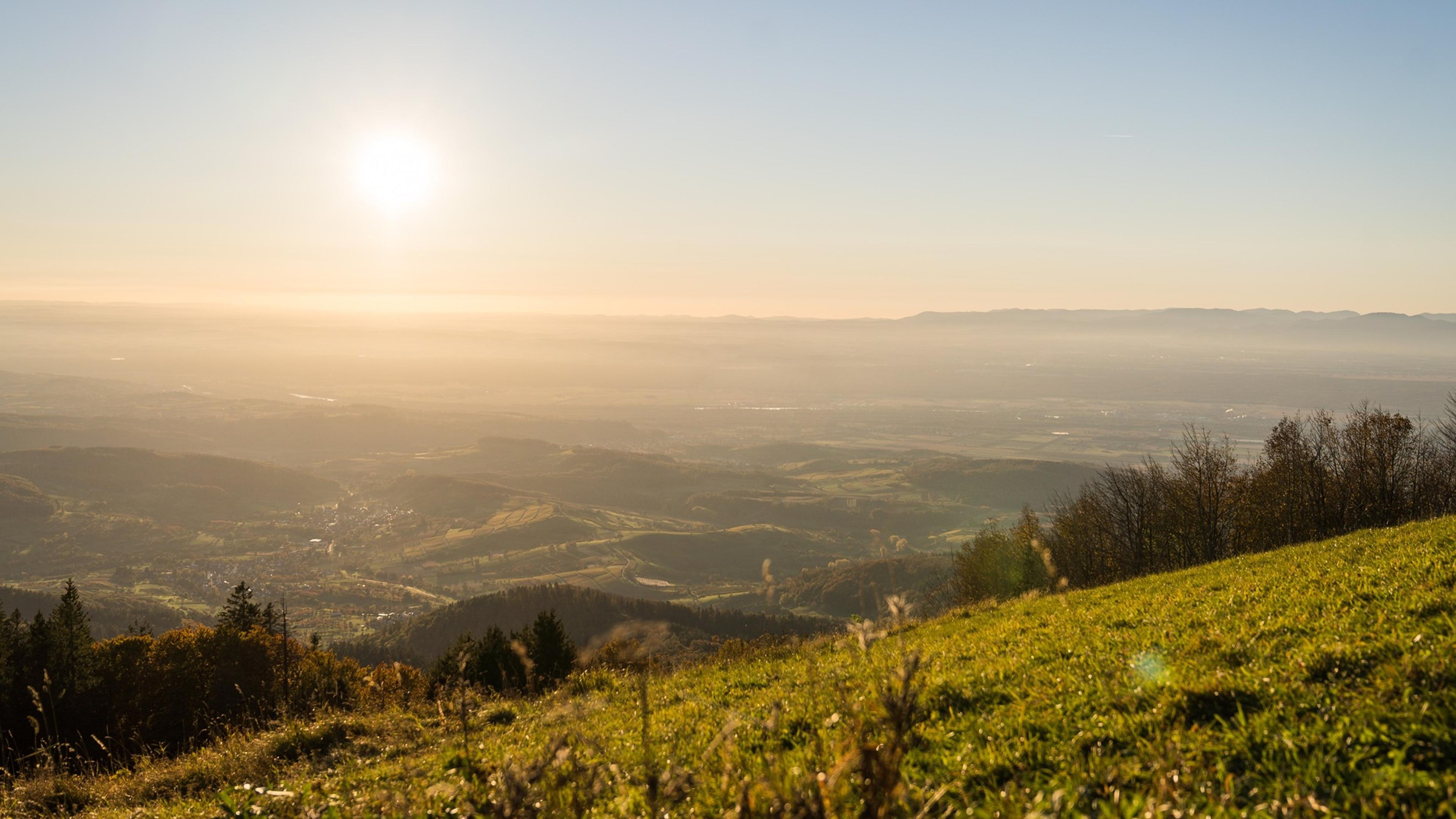 Sunrise over a vast landscape with rolling hills, grassy fields, distant mountains, and a clear sky.