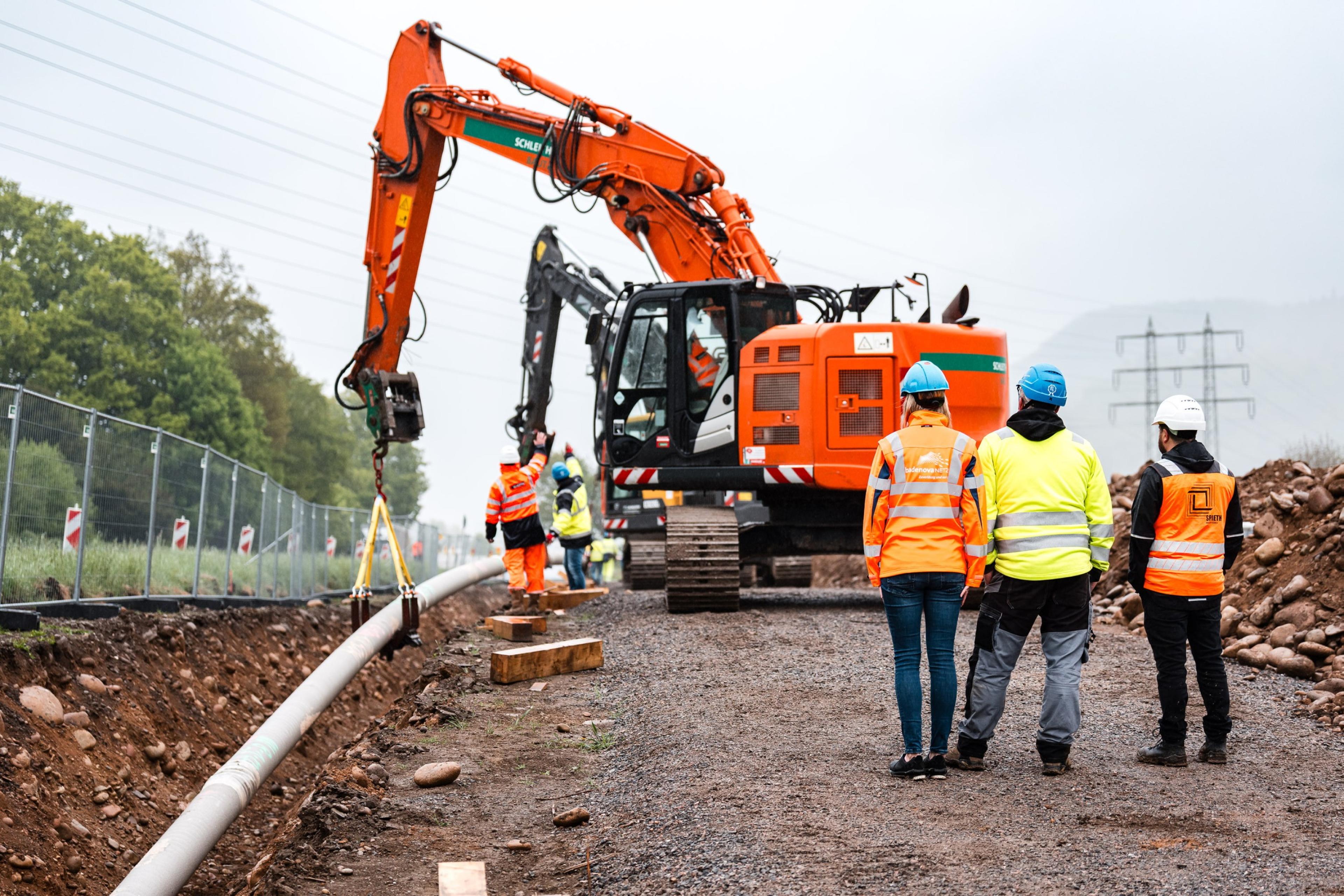 Bauarbeiter von BadenovaNETZE und ein großer Bagger verlegen eine Rohrleitung in einer ländlichen Gegend. Die Arbeiter tragen Schutzkleidung, und der Himmel ist bewölkt.