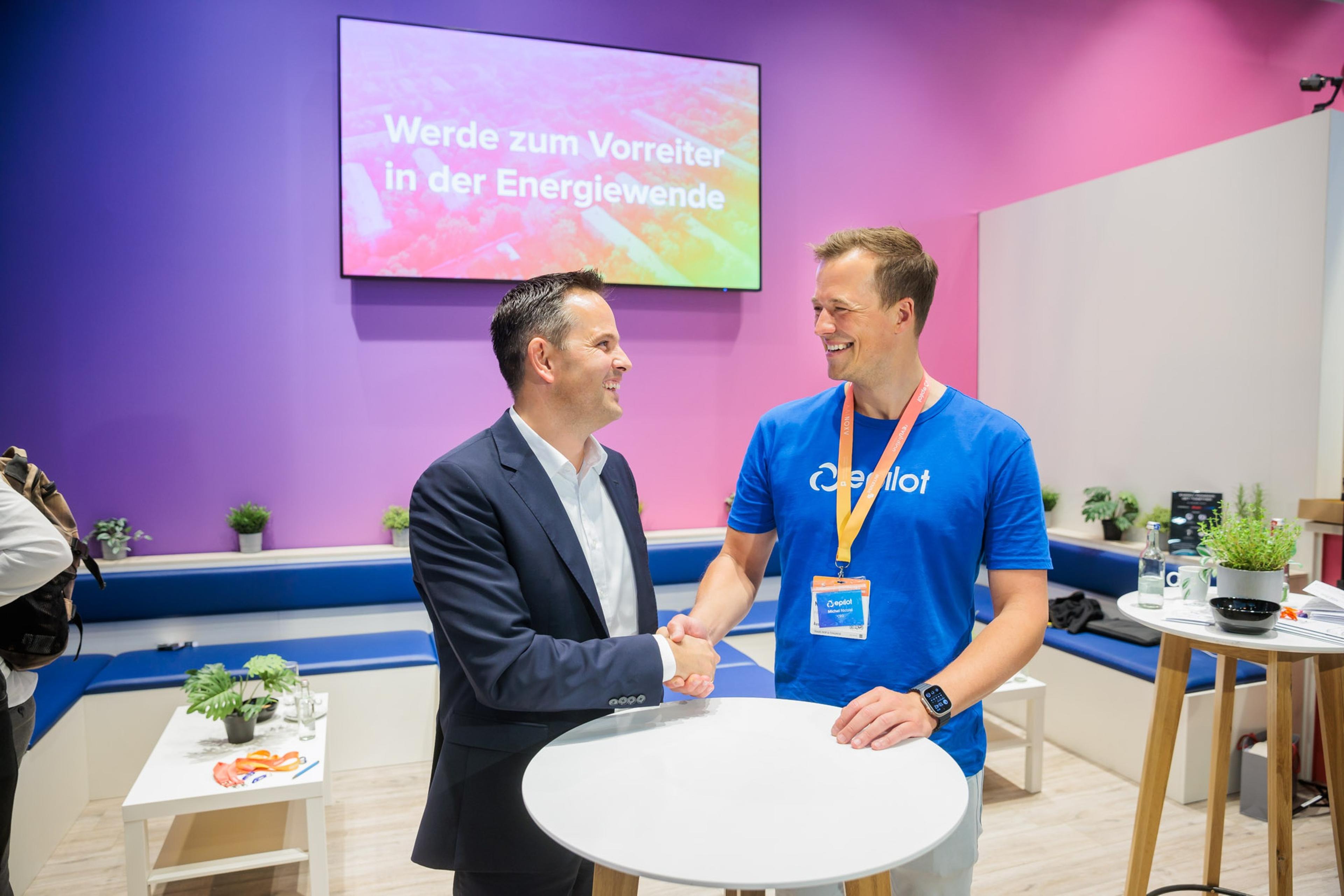 Zwei Männer geben sich in einer Kabine mit rosa und violetter Wand die Hand. Auf einem Bildschirm wird deutscher Text angezeigt. Auf den Tischen stehen Pflanzen und Snacks.