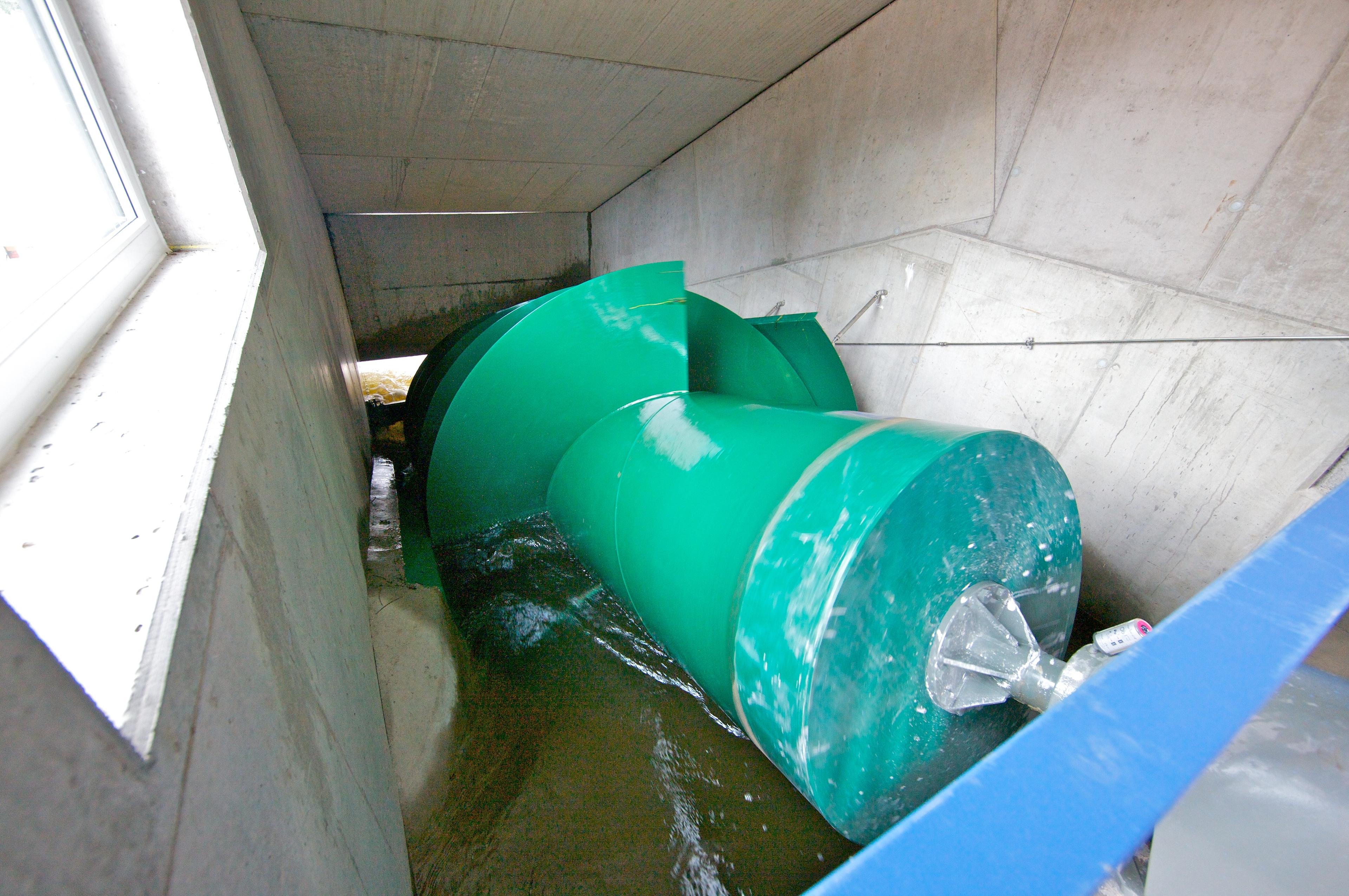 A large green screw turbine is installed in a concrete channel with flowing water in a hydroelectric power plant setting.