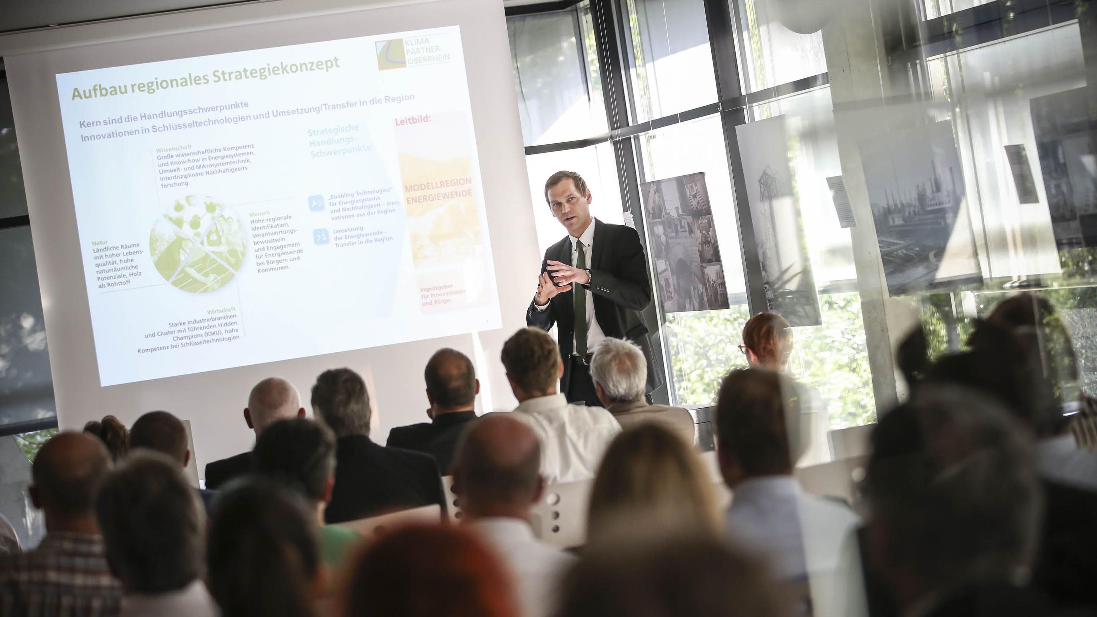 Ein Mann im Anzug stellt einem Publikum ein strategisches Konzept vor, während in einem hellen Konferenzraum Folien auf einer Leinwand gezeigt werden.