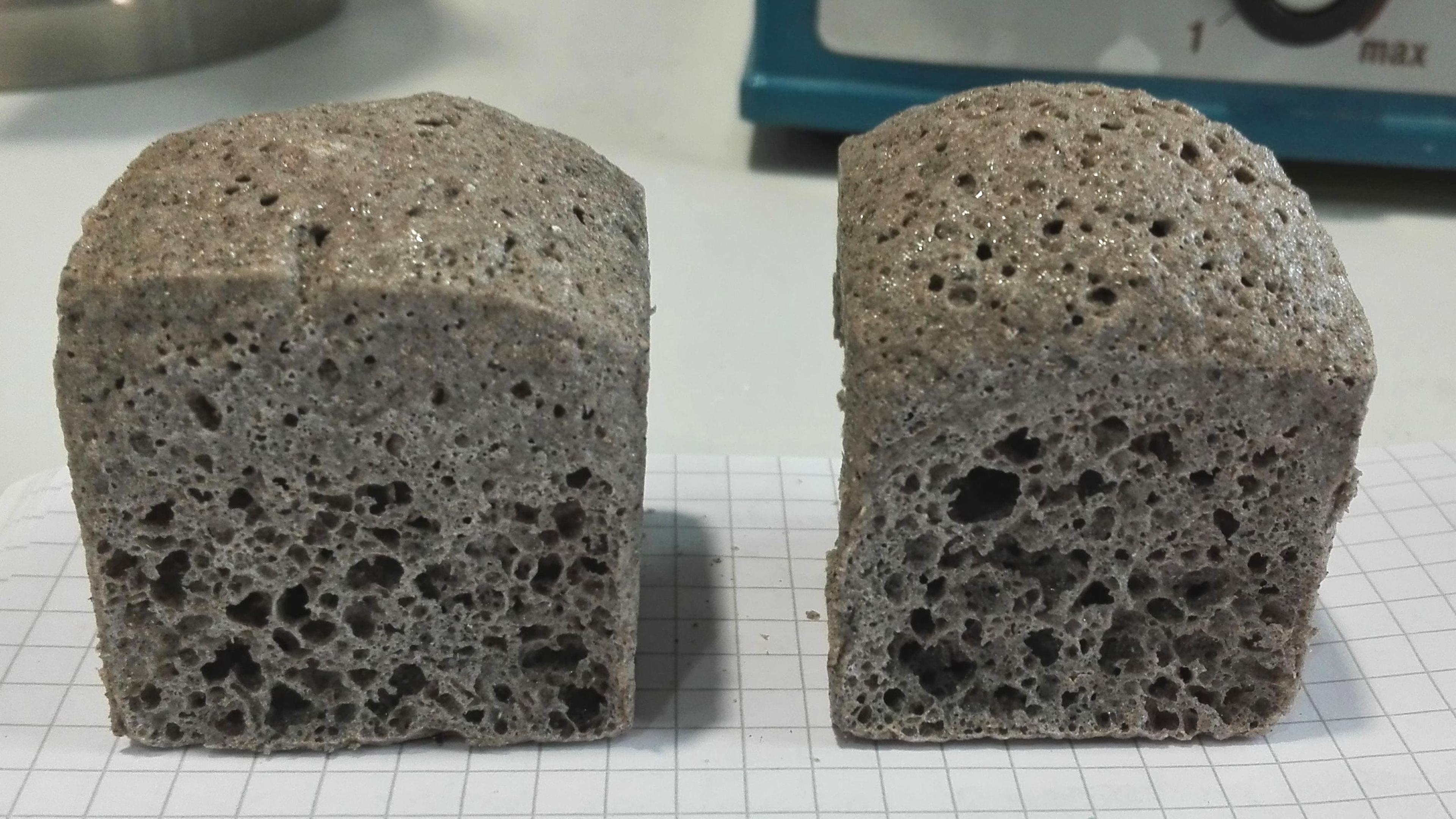 Zwei poröse, graue Betonwürfel auf Millimeterpapier, die ihre strukturierten Oberflächen und kleinen Luftlöcher zeigen.