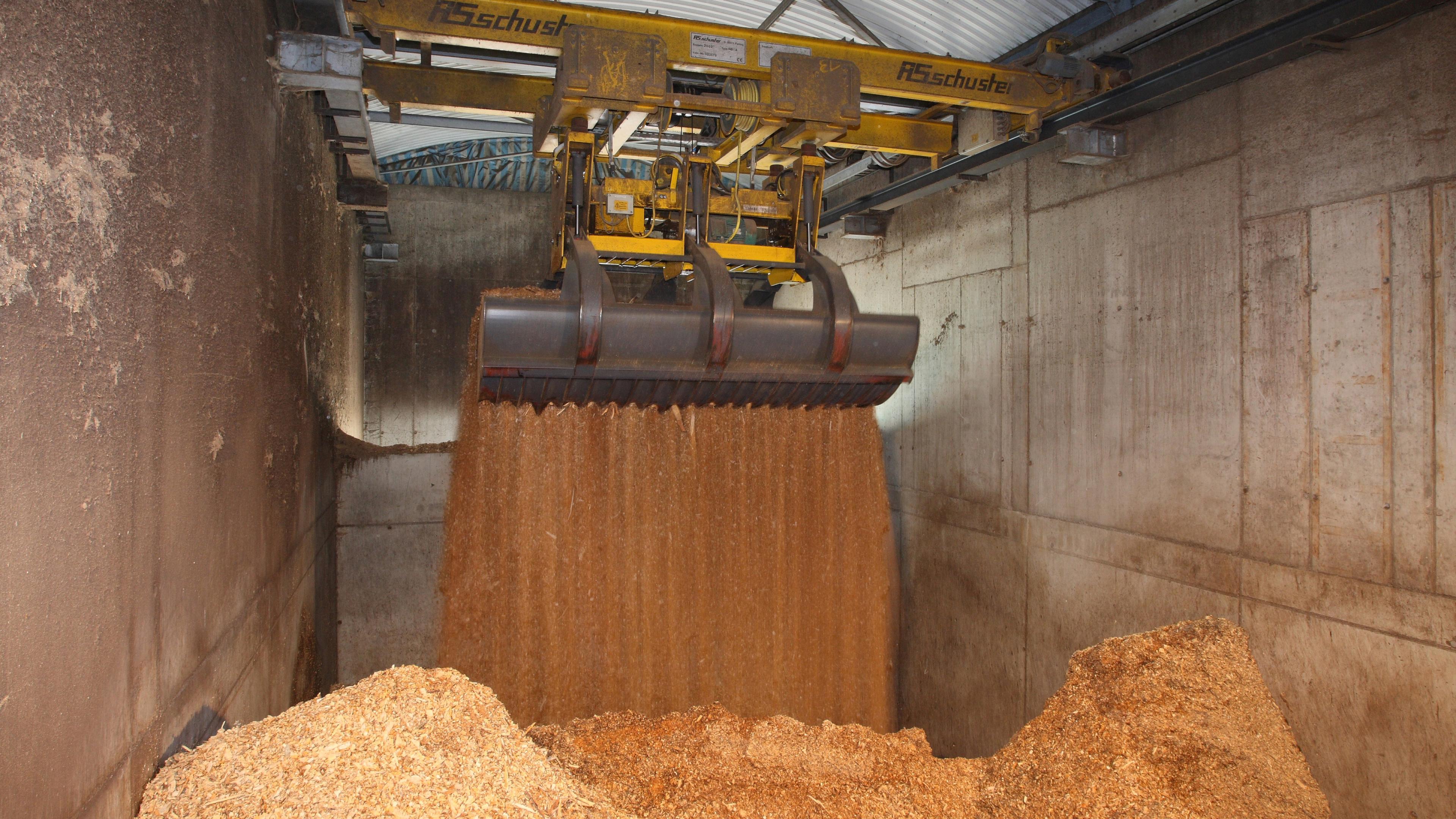 Ein großer Industriegreifer schüttet einen Strom von Holzspänen in einen Lagerbereich, wo sich die Späne unten auf dem Betonboden zu Haufen ansammeln.