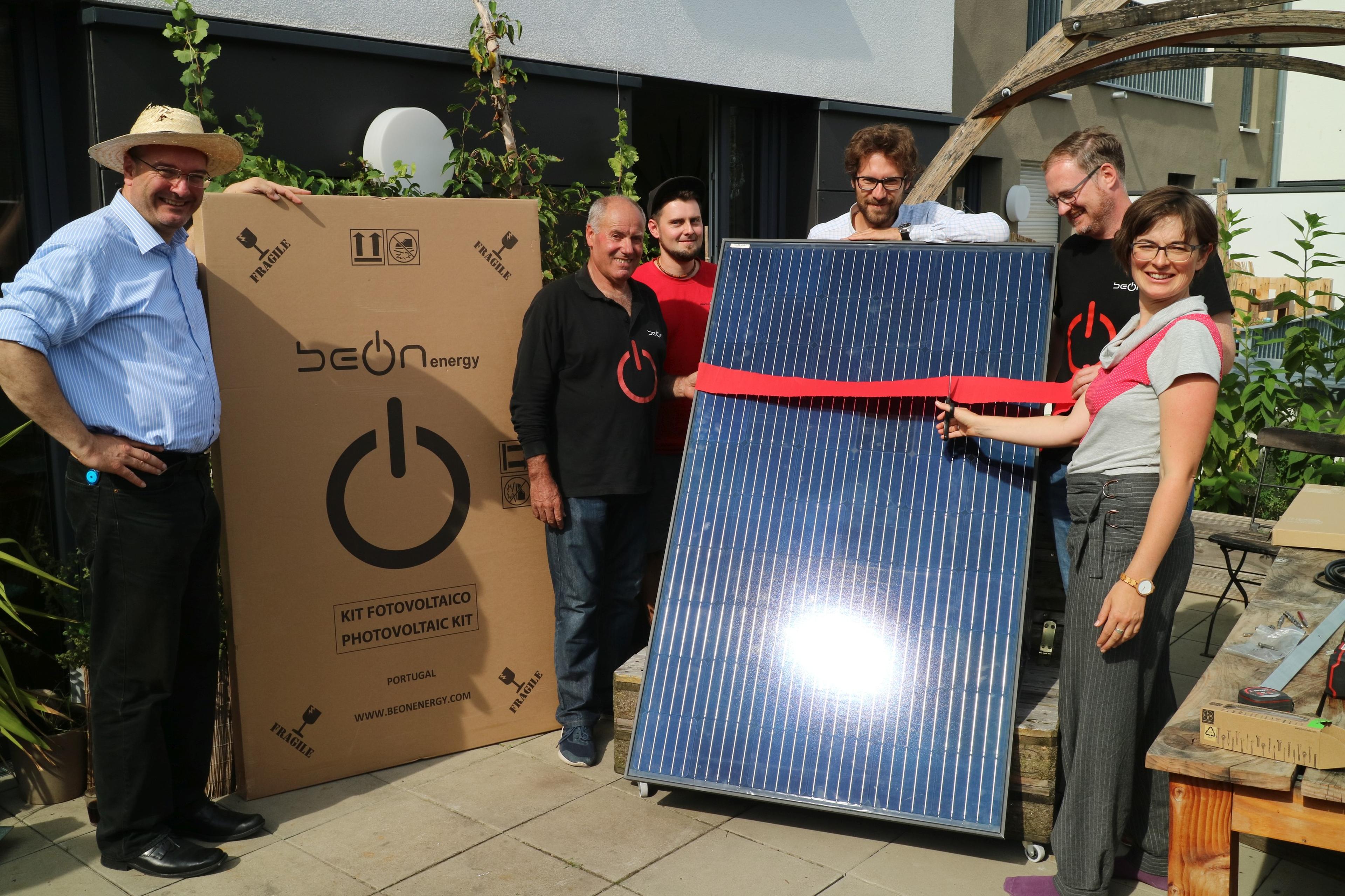 Eine Gruppe von Menschen lächelt und hält ein großes Solarpanel auf einer sonnigen Terrasse.