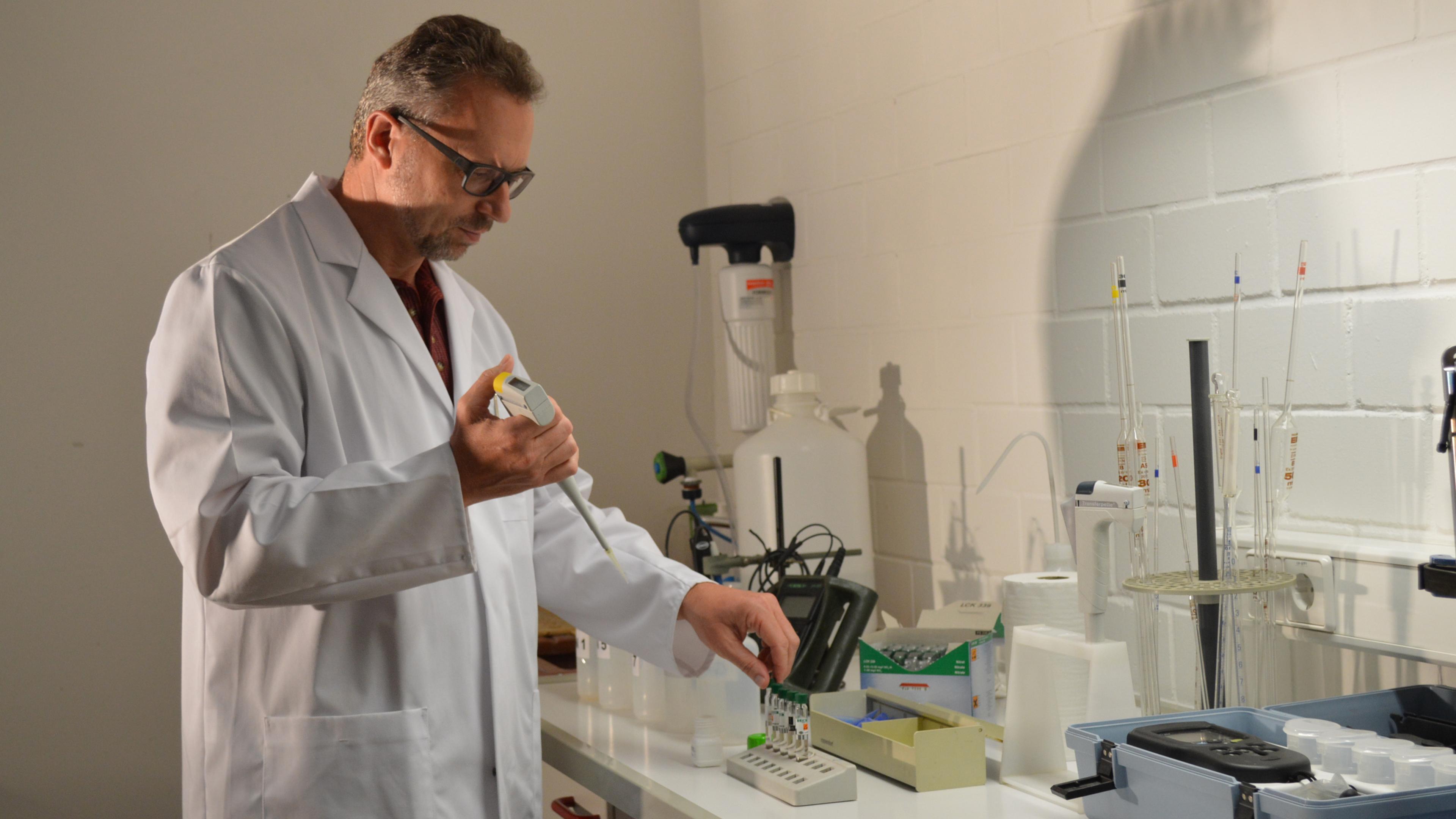 Ein Wissenschaftler im Laborkittel bereitet an einem Labortisch, umgeben von Laborgeräten, mit einer Pipette Proben vor.