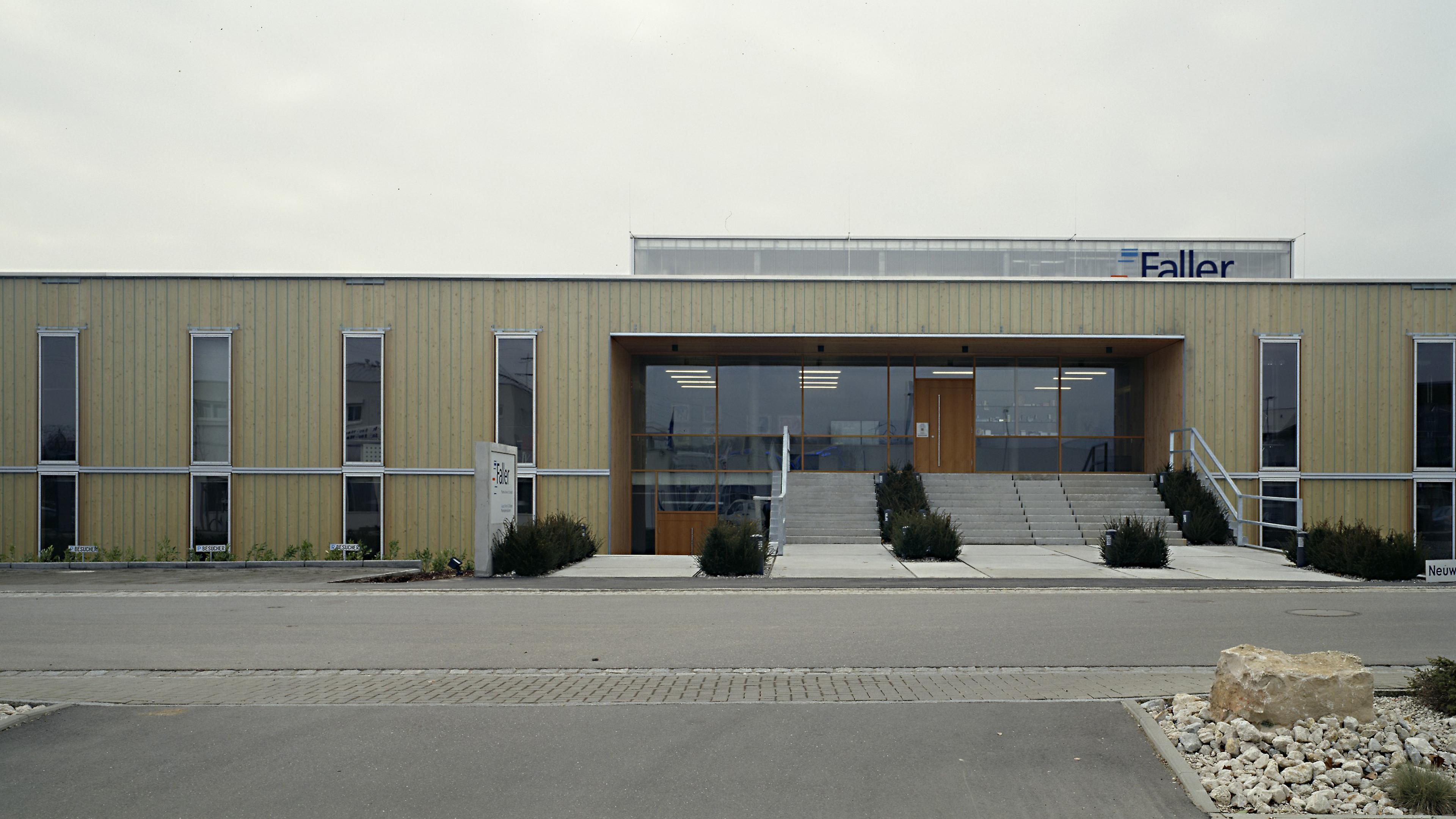 Vorderansicht eines modernen, beigen Gebäudes mit großen Fenstern, einem zentralen Eingang mit Treppe und einem Schild mit dem Namen „Faller“, das an der Fassade zu sehen ist.