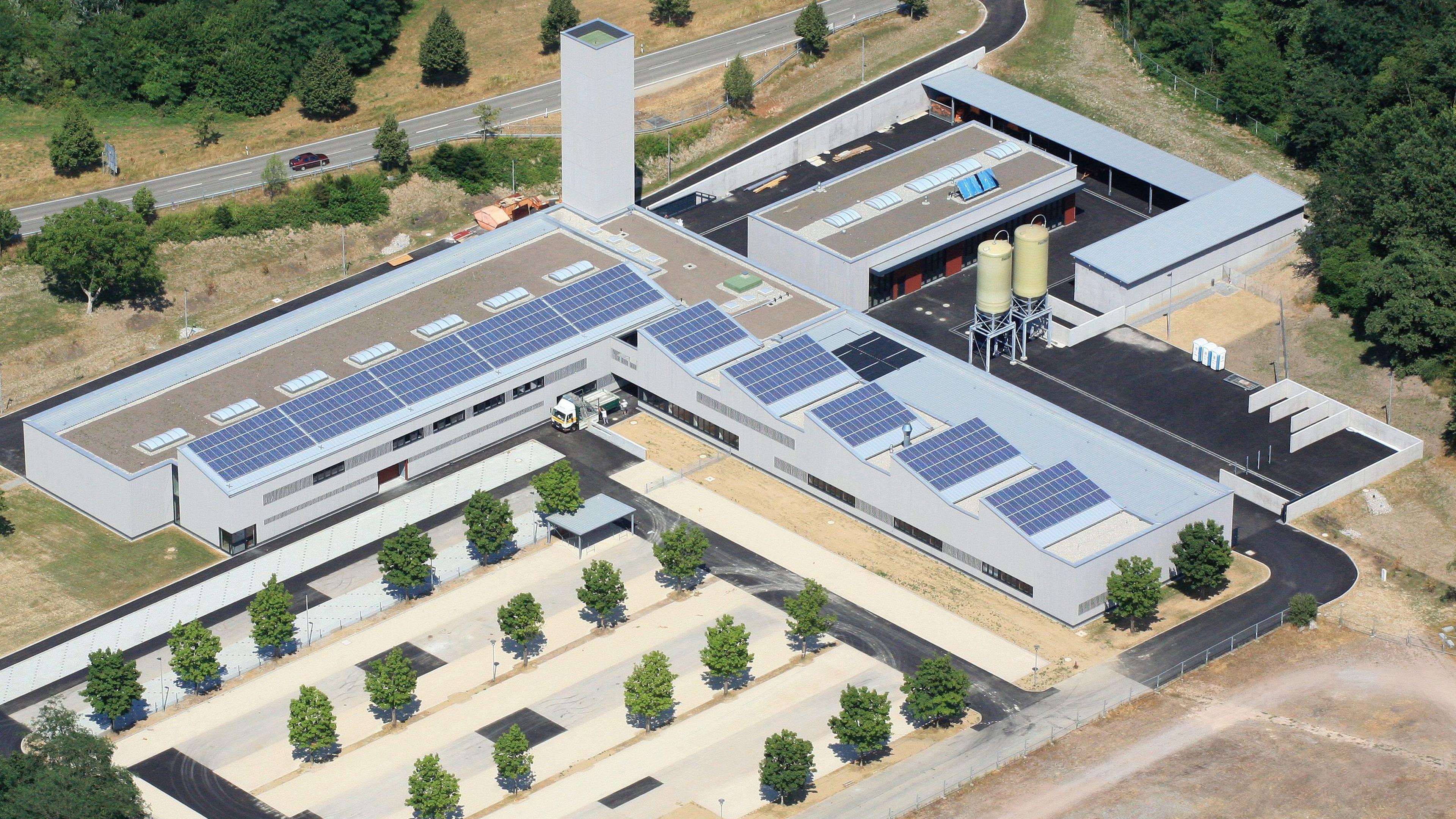 Luftaufnahme einer Industrieanlage mit Solarmodulen auf den Daechern, umgeben von Baeumen und einer Strasse im Hintergrund.