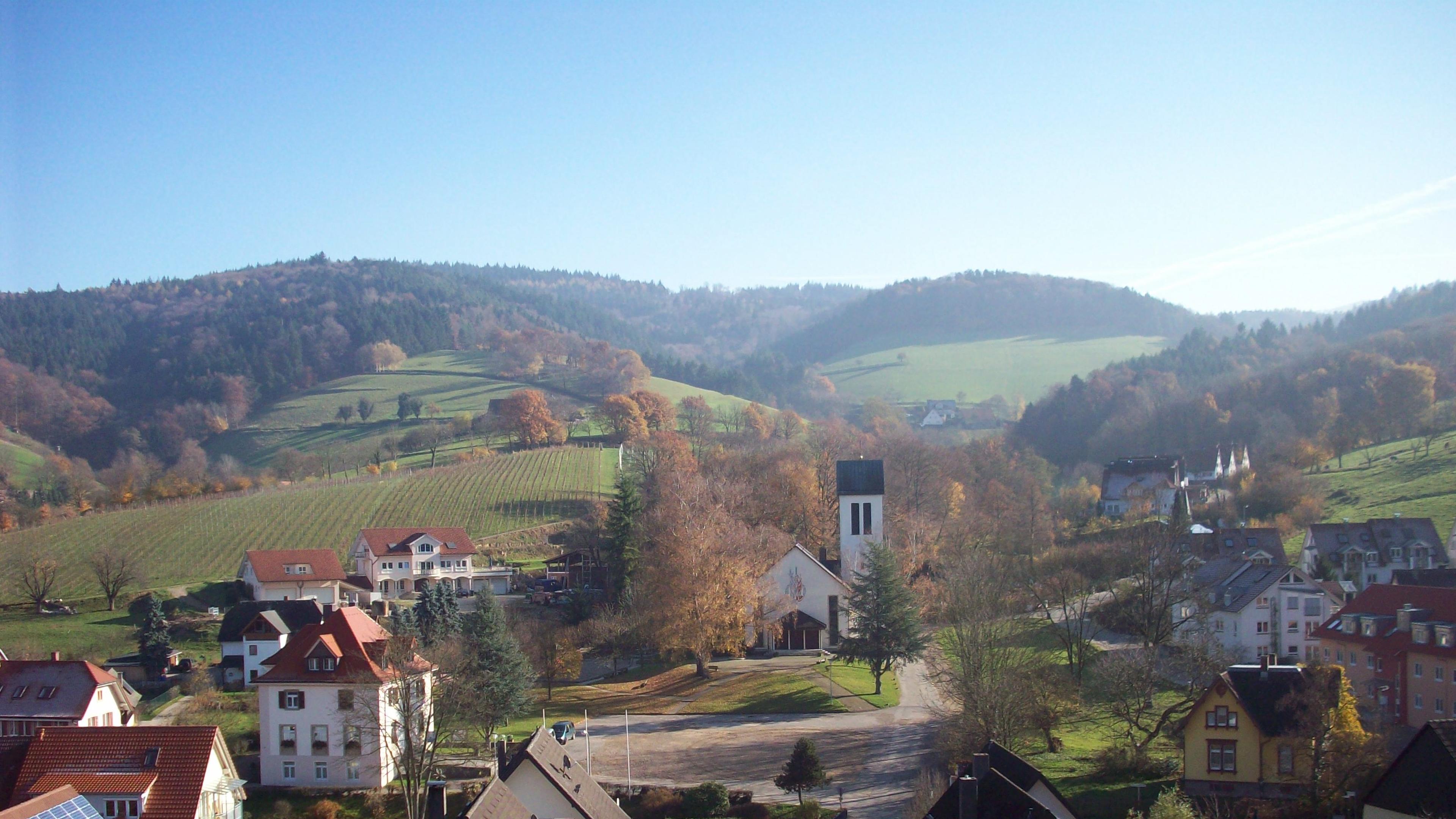 Ein malerisches Dorf mit einer Kirche, umgeben von sanften Hügeln, Weinbergen und herbstlich gefärbten Bäumen unter strahlend blauem Himmel.
