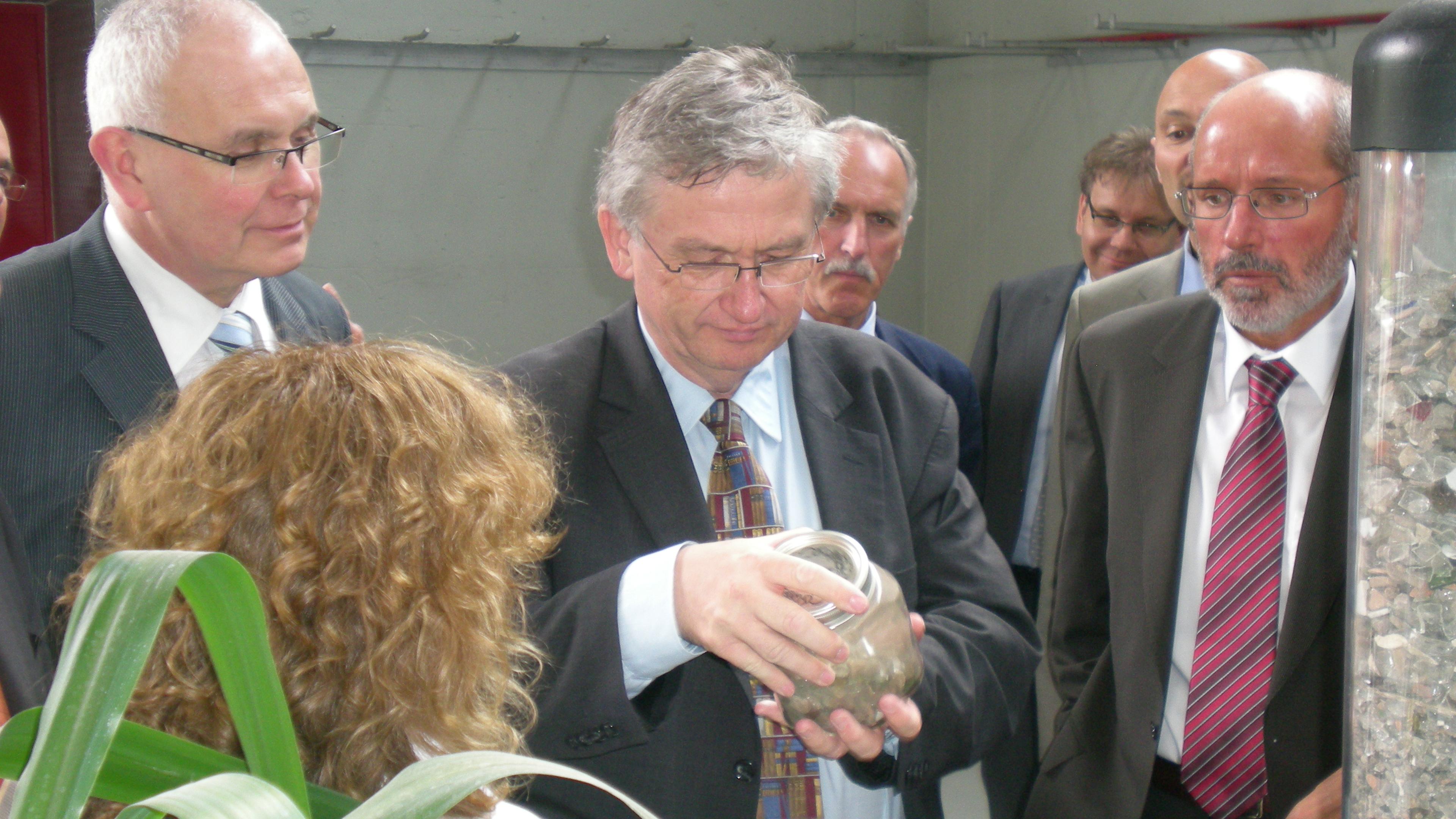Ein Mann im Anzug begutachtet bei einer Veranstaltung in einem Saal ein Glas mit Materialien, umgeben von anderen Männern in Anzügen.