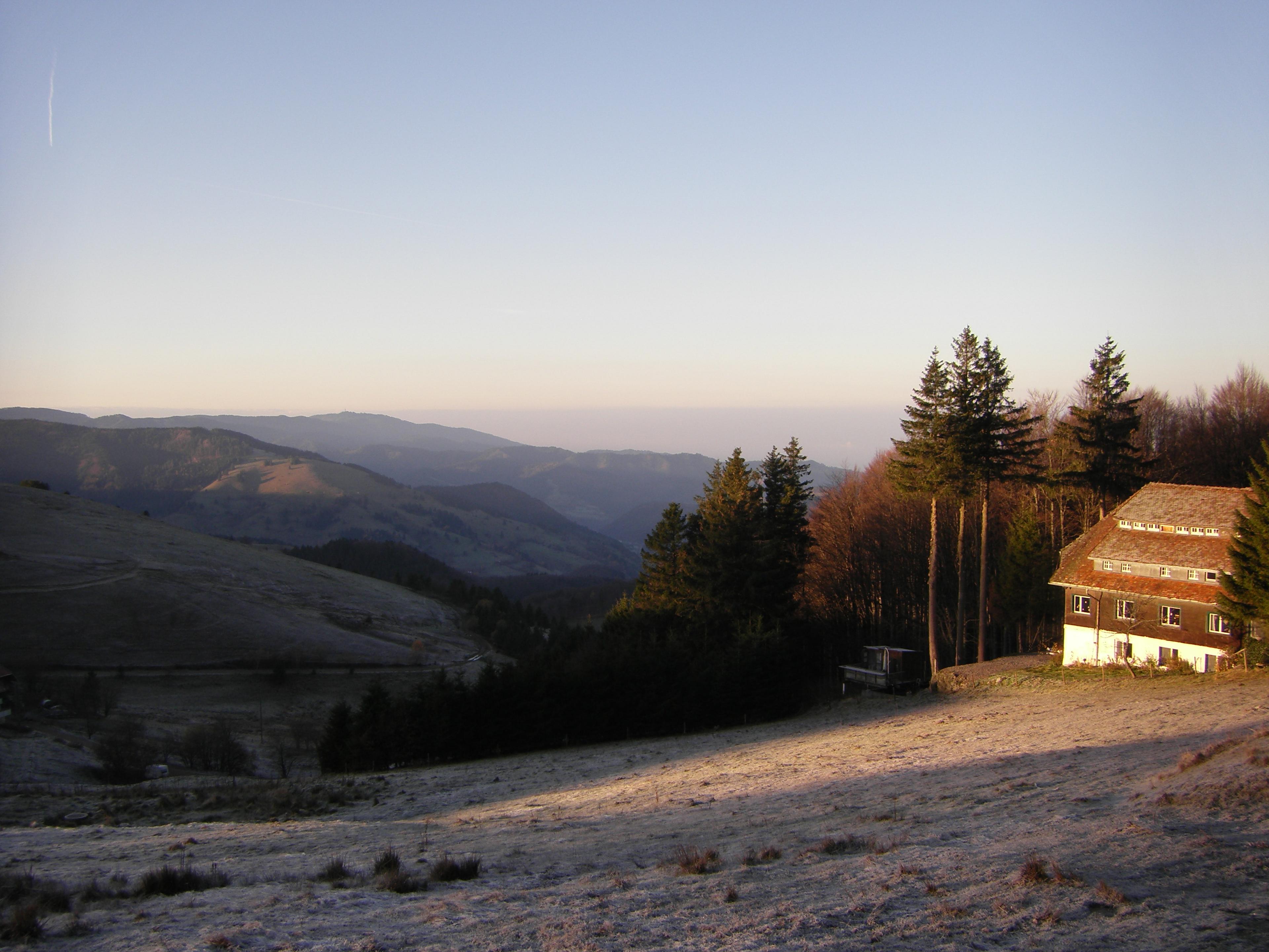 Malerischer Blick auf einen frostbedeckten Hang mit einem rustikalen Haus, umgeben von hohen Bäumen, unter klarem Himmel und mit fernen Bergketten im Hintergrund bei Sonnenuntergang.