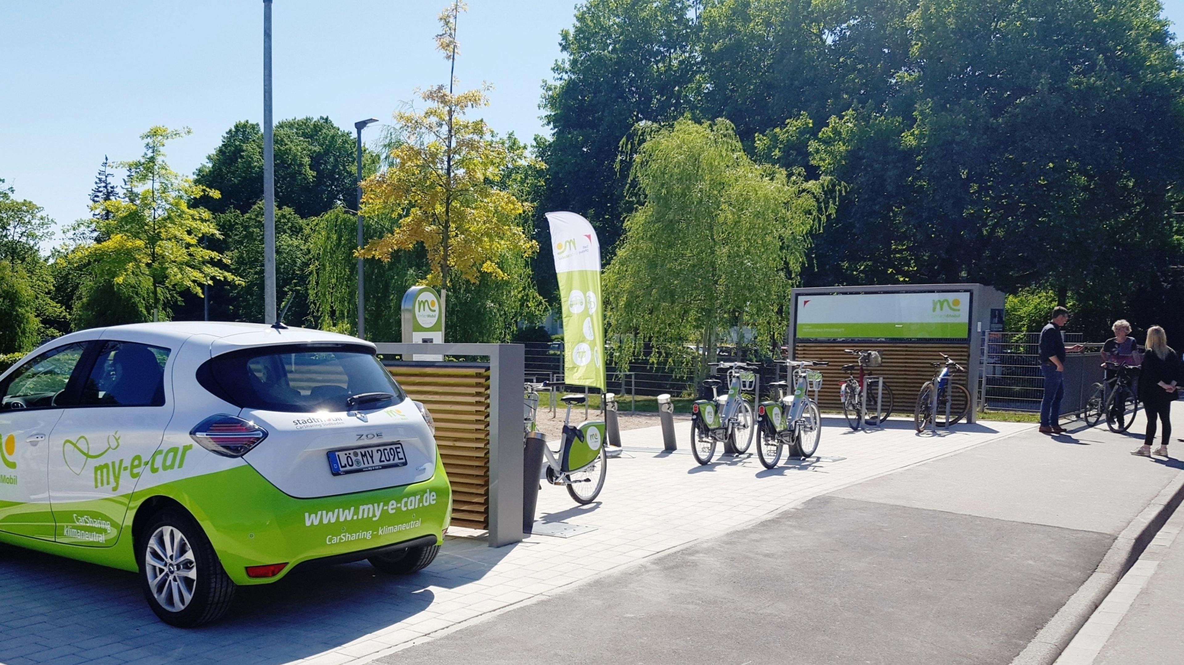 Elektroautos und -fahrräder an einer Ladestation mit grünem Branding. Menschen in der Nähe und Bäume im Hintergrund unter klarem Himmel.