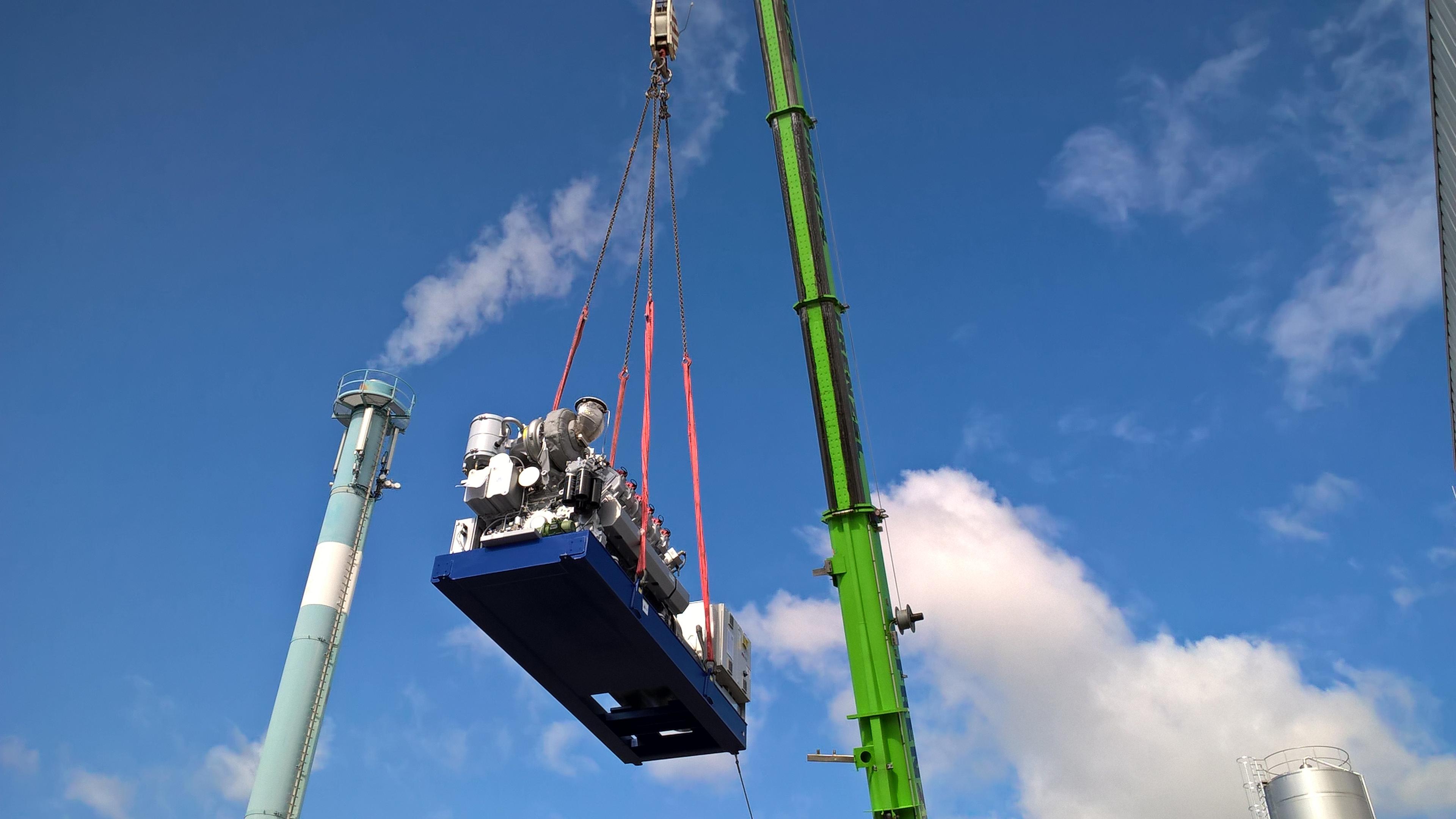 Eine große Maschine wird von einem grünen Kran vor einem blauen Himmel mit Wolken in der Nähe von Industrieschornsteinen angehoben.