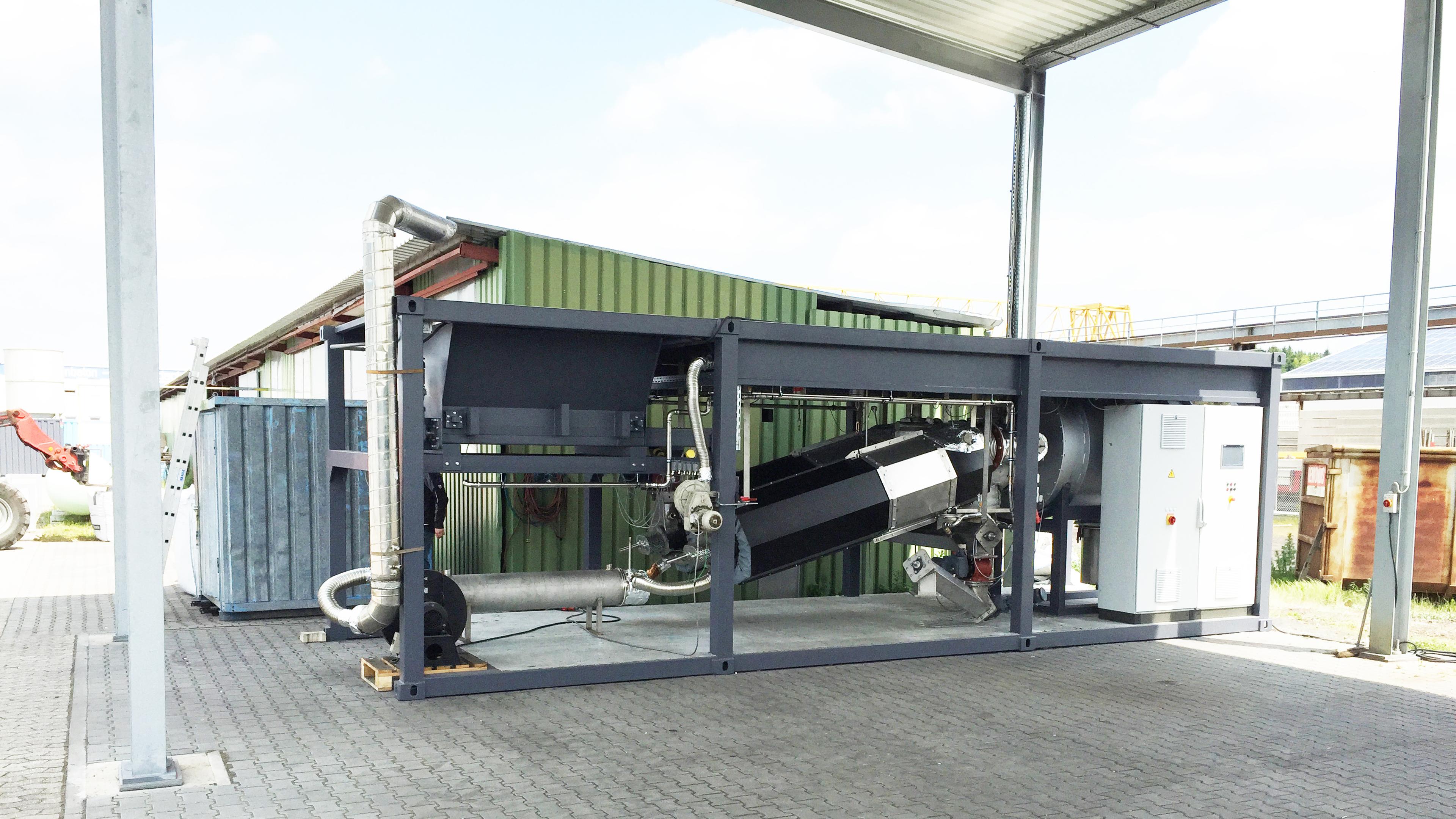 Industrial machinery setup outdoors under a metal shelter, with a green building in the background on a paved surface.