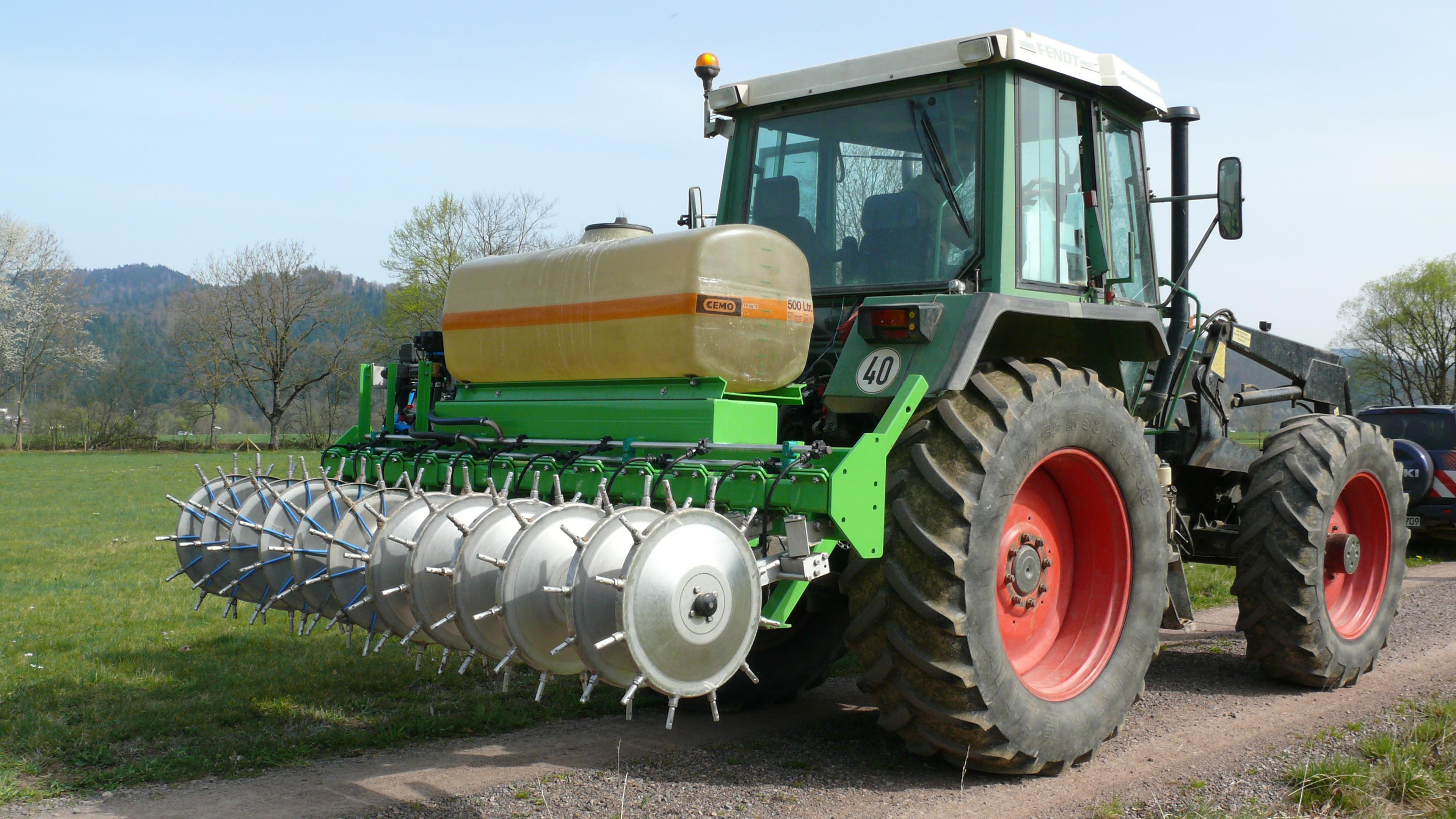 Ein Traktor mit großen roten Rädern und einem grünen Anbaugerät auf einem Feldweg, vor dem Hintergrund von Bäumen und Hügeln.