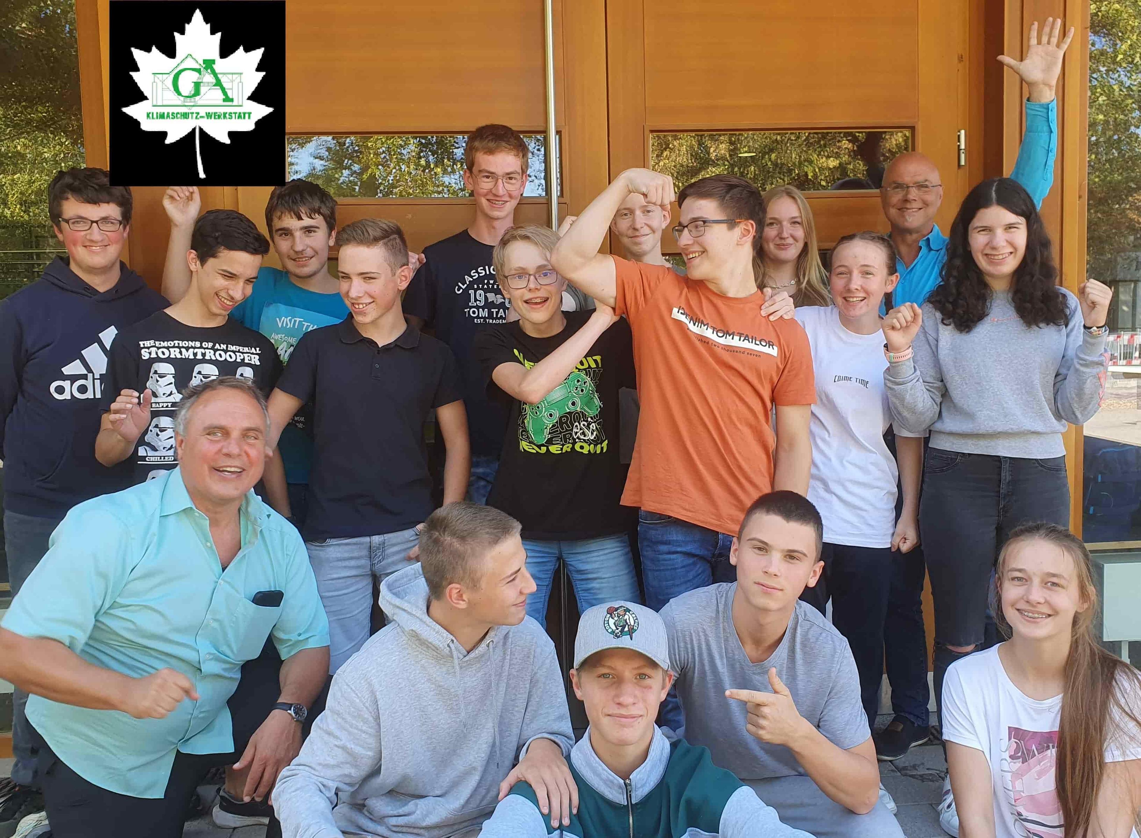 Eine Gruppe lächelnder Menschen, von denen einige zur Feier ihre Arme hochreißen, posiert vor einer Holztür. In der Ecke befindet sich ein Logo mit einem Ahornblatt.