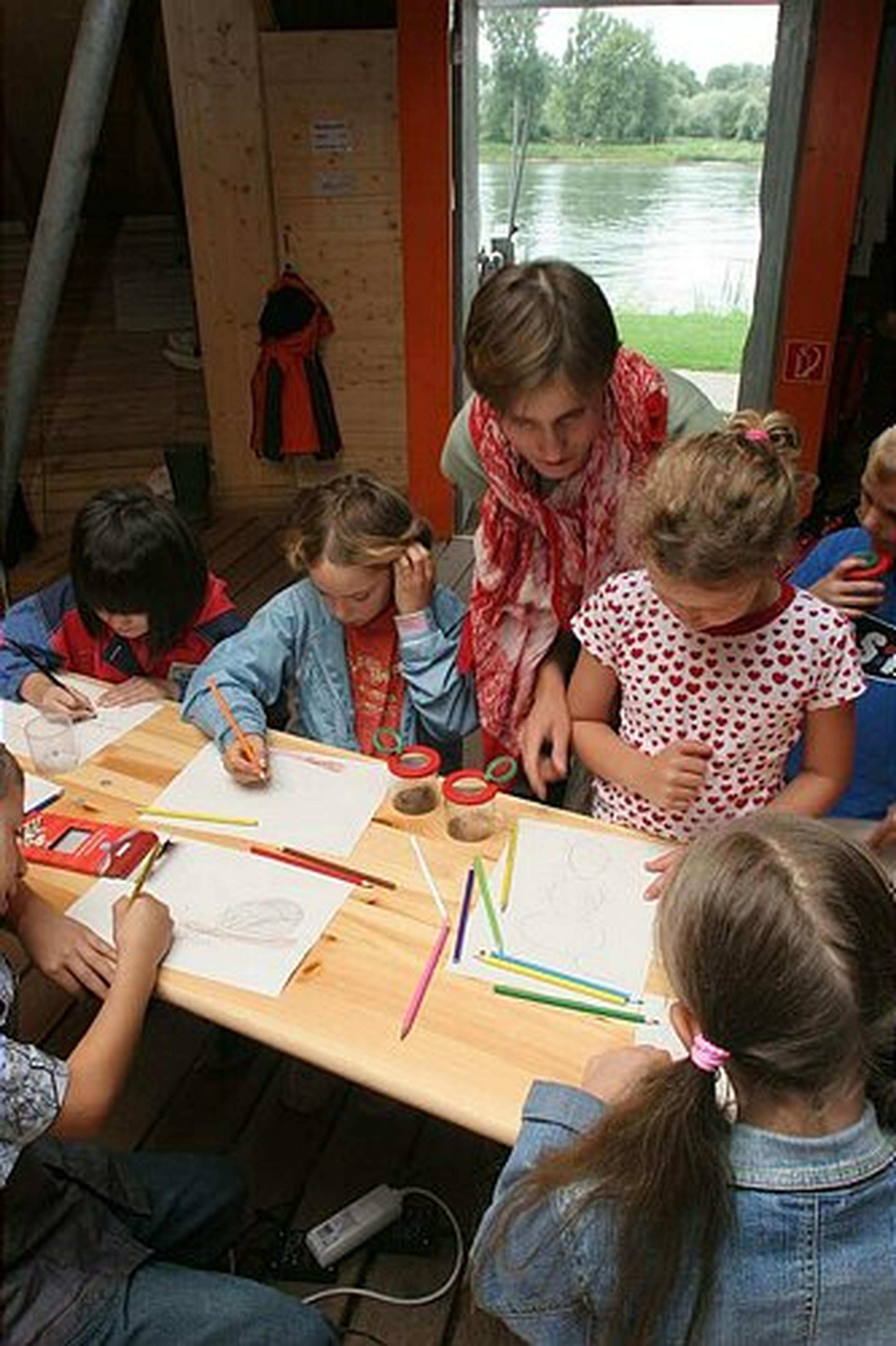 Kinder, die an einem Tisch mit Buntstiften malen, angeleitet von einem Erwachsenen. Sie befinden sich in einem gemütlichen Raum mit Blick auf Bäume und einen See draußen.
