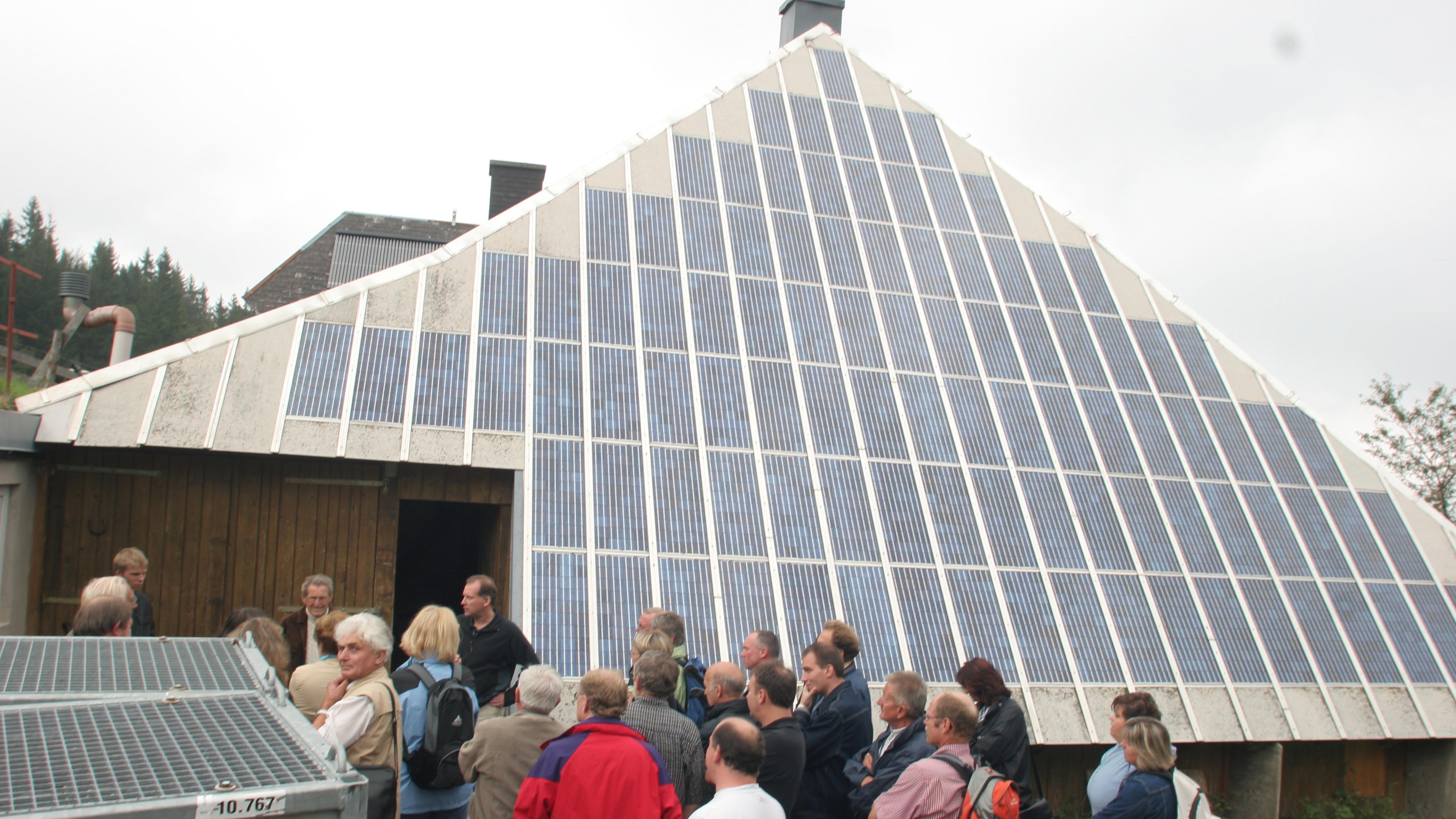 Eine Gruppe von Menschen steht an einem bewölkten Tag vor einem Gebäude mit einer großen dreieckigen Solarfassade, das von Bäumen umgeben ist.