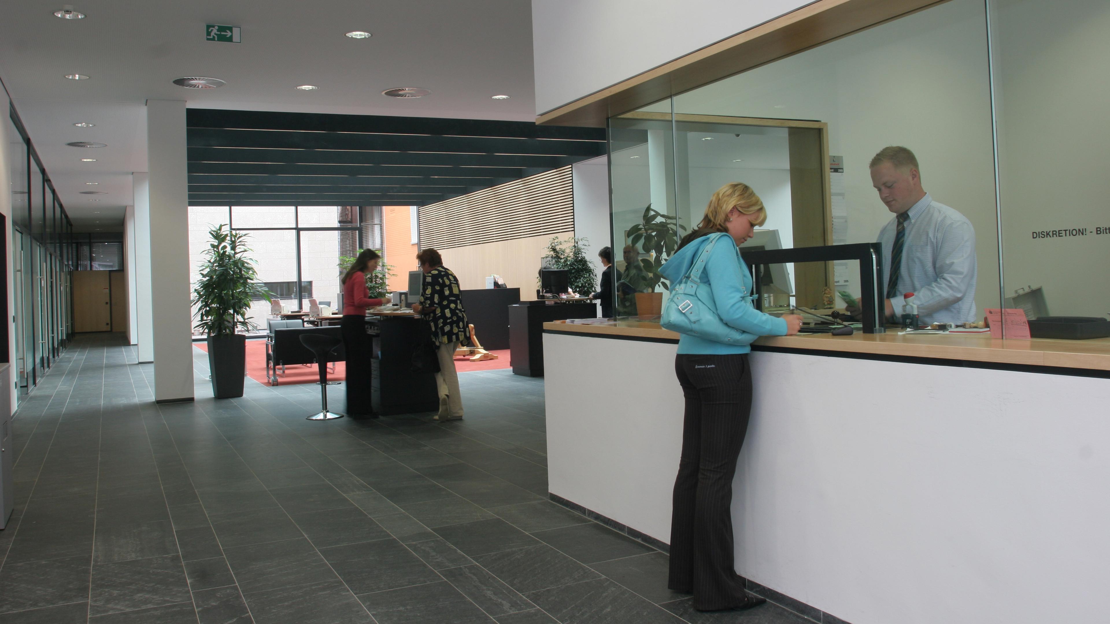 Eine moderne Büro-Lobby, in der sich Menschen an einem Empfangstresen und an Arbeitsplätzen unterhalten. Im Hintergrund sind Pflanzen und ein Flur zu sehen.