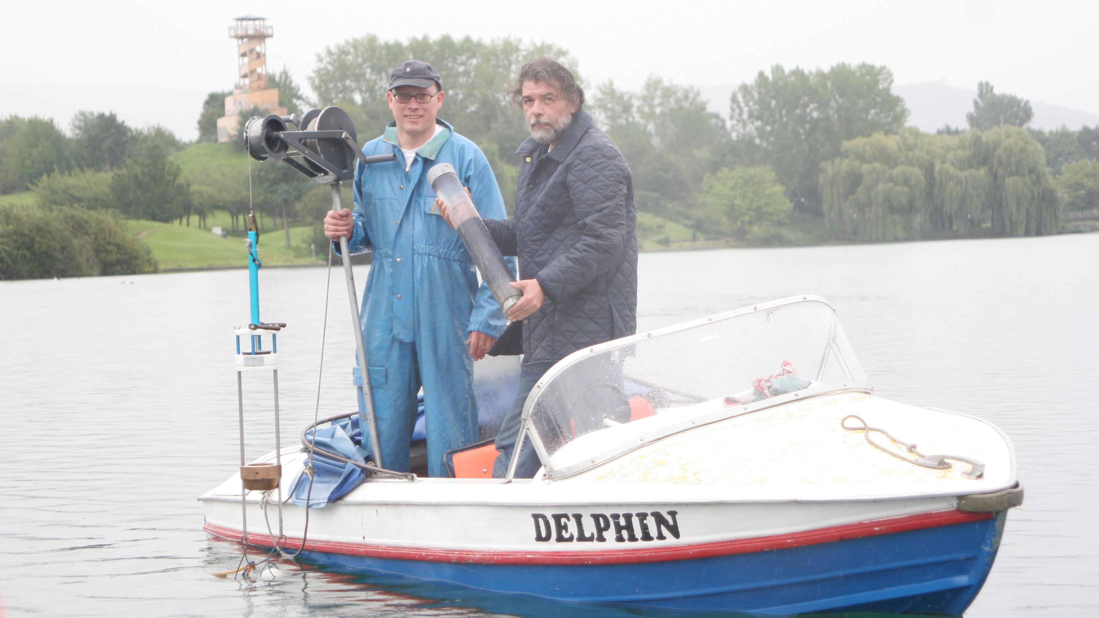 Zwei Männer auf einem kleinen Boot namens „Delphin“ auf einem See, die wissenschaftliche Geräte in den Händen halten. Im Hintergrund sind ein Turm und Bäume zu sehen.