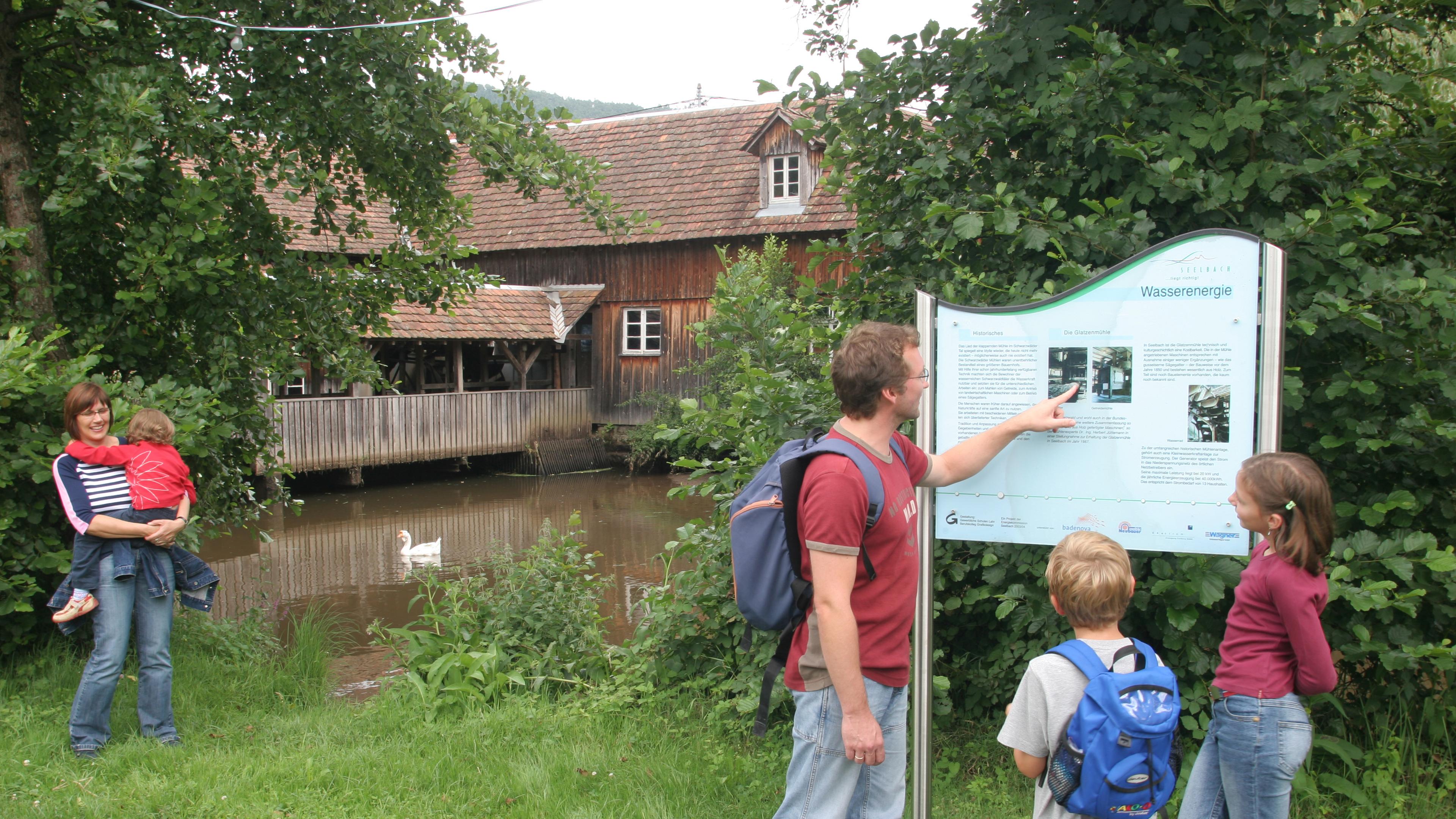 Eine Familie betrachtet eine Informationstafel zum Thema Wasserkraft in der Nähe eines rustikalen Gebäudes an einem Teich mit einem Schwan, umgeben von üppigem Grün.