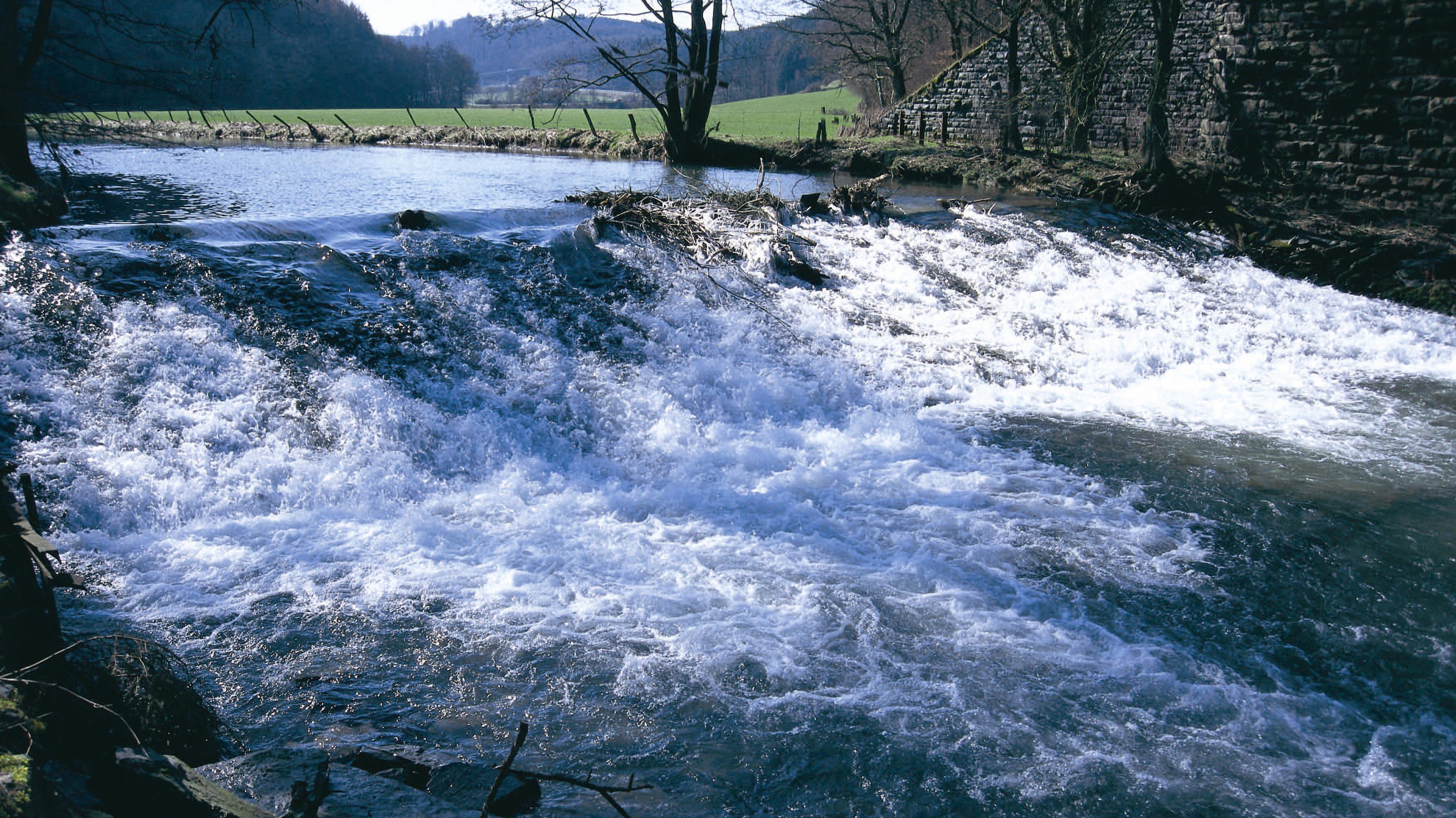 Ein kleiner Wasserfall, der über Felsen in einen Fluss stürzt, umgeben von Bäumen und einer Steinmauer unter einem klaren Himmel.