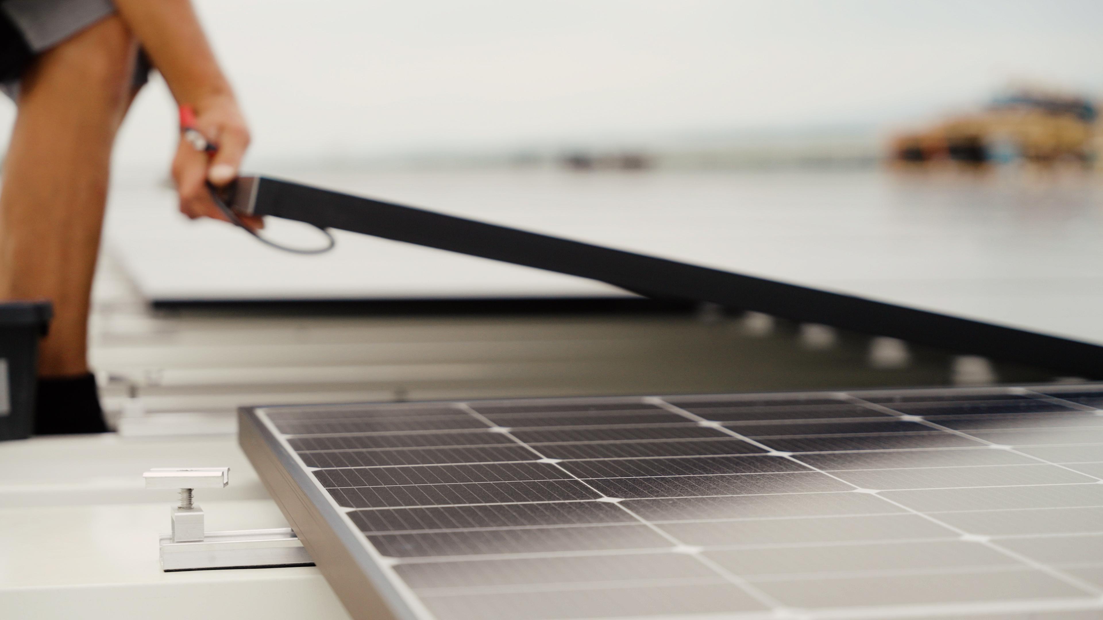 Eine Person, die auf einem Dach Solarmodule installiert, mit Fokus auf eine Hand, die ein Modul ausrichtet. Im Hintergrund ein strahlend blauer Himmel.