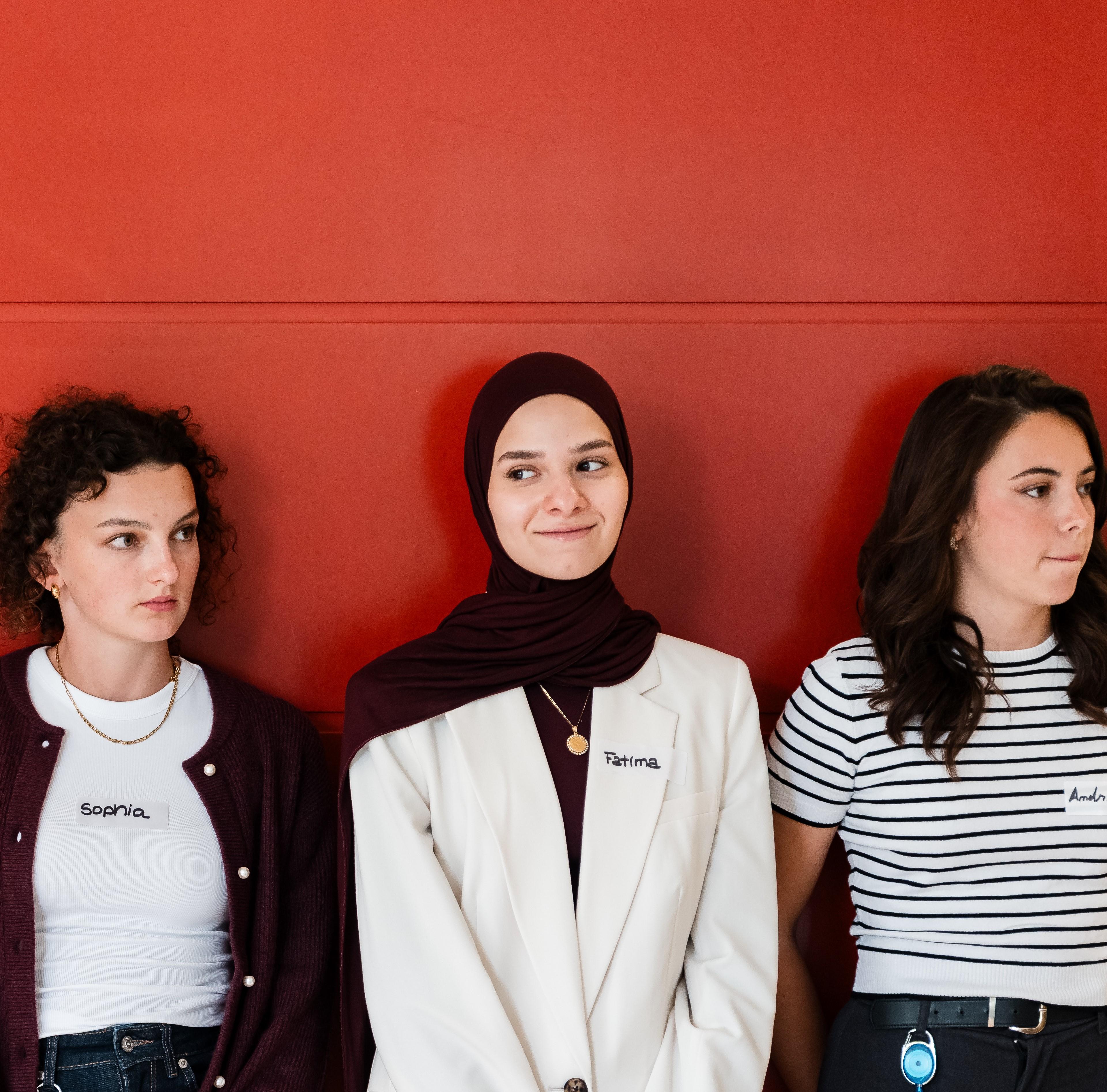 Drei Frauen stehen vor einer roten Wand, jede mit einem Namensschild. Die Frau in der Mitte trägt einen Hidschab und einen weißen Blazer.