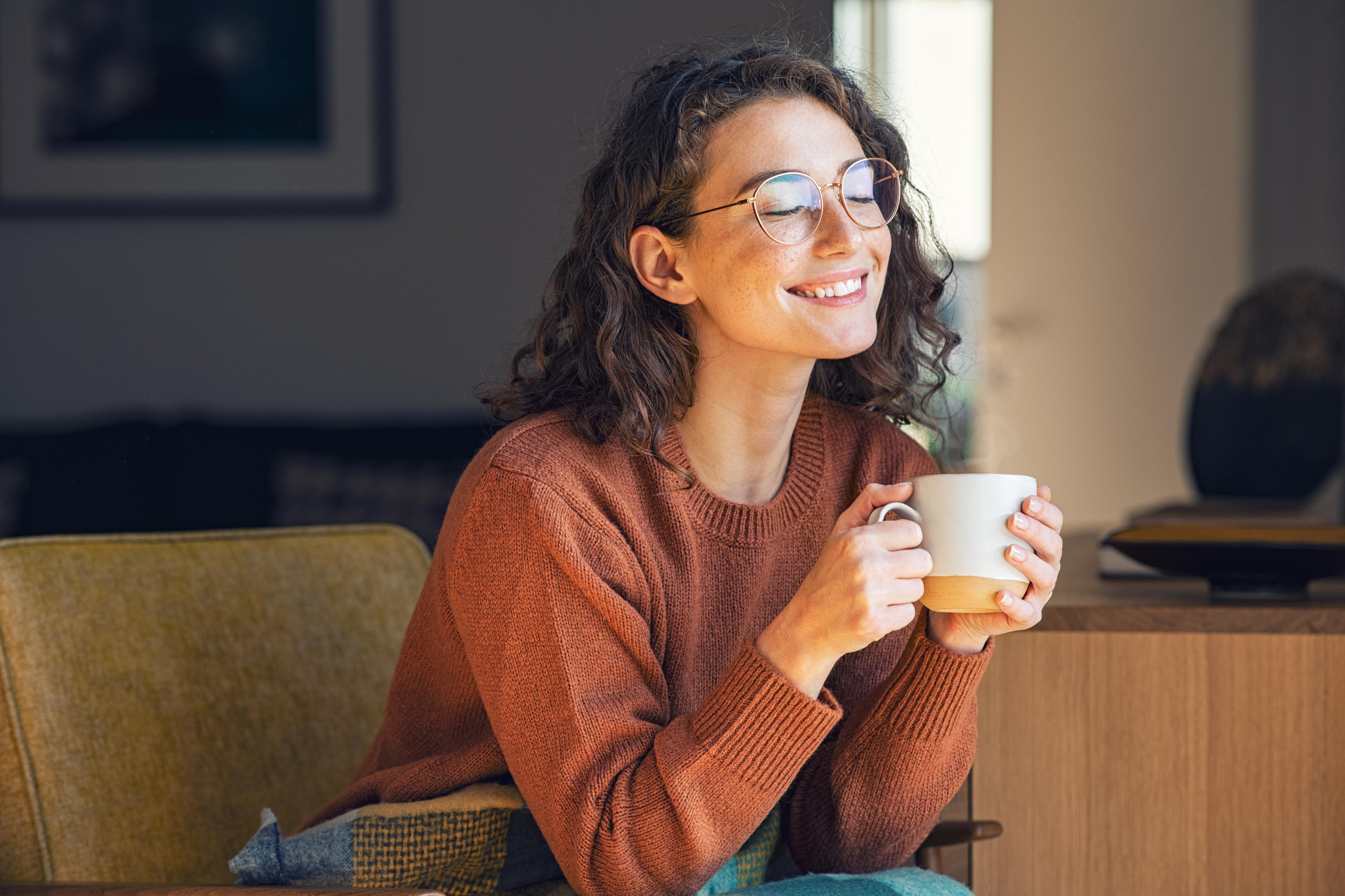 Lächelnde Frau mit lockigem Haar und Brille, die eine Kaffeetasse hält und auf einer Couch in einem gemütlichen Zimmer sitzt.