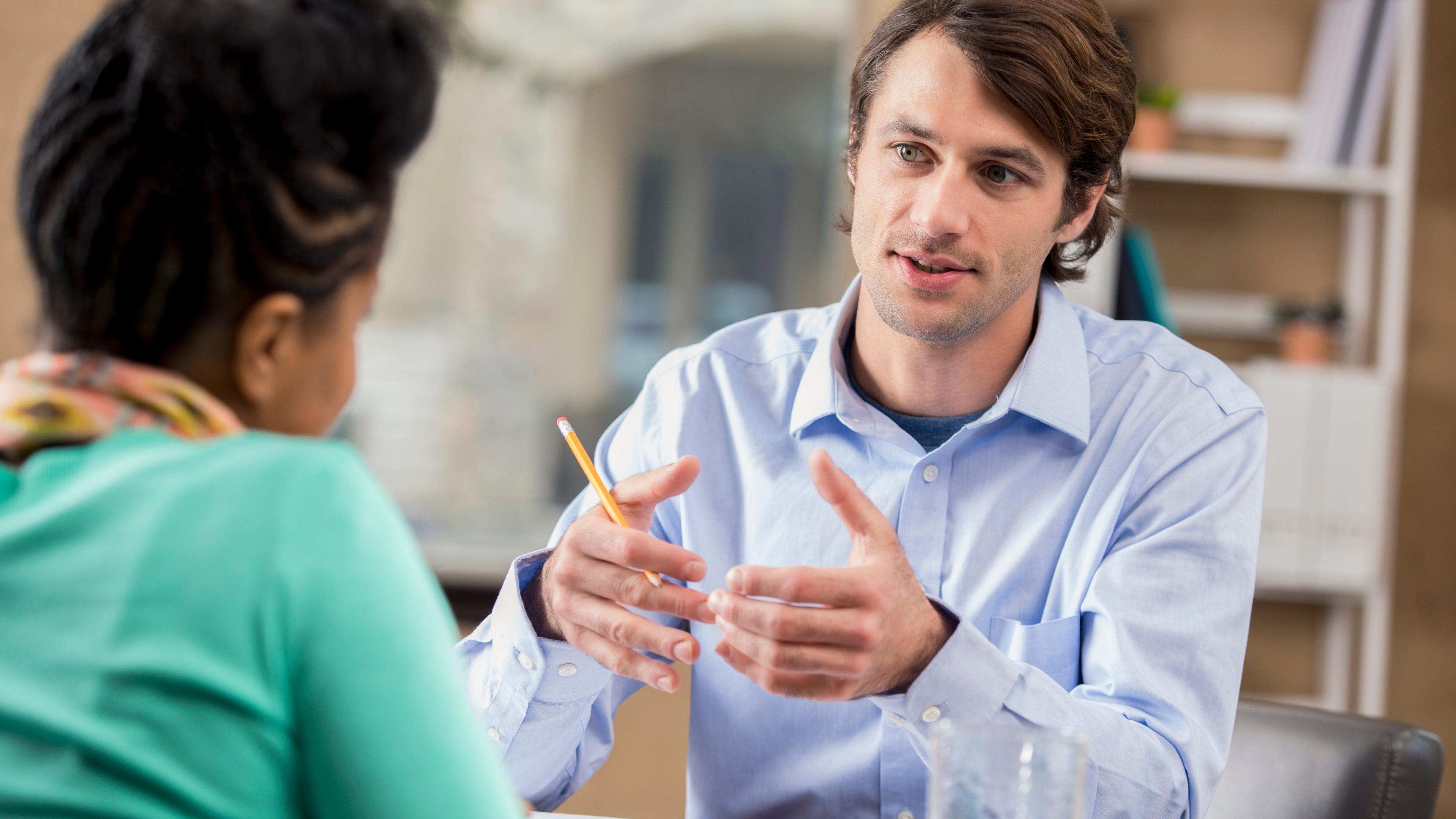Mann in einem hellblauen Hemd gestikuliert mit einem Bleistift, während er mit einer Person spricht, die ihm gegenüber an einem Tisch mit Notizbüchern und einem Glas sitzt.