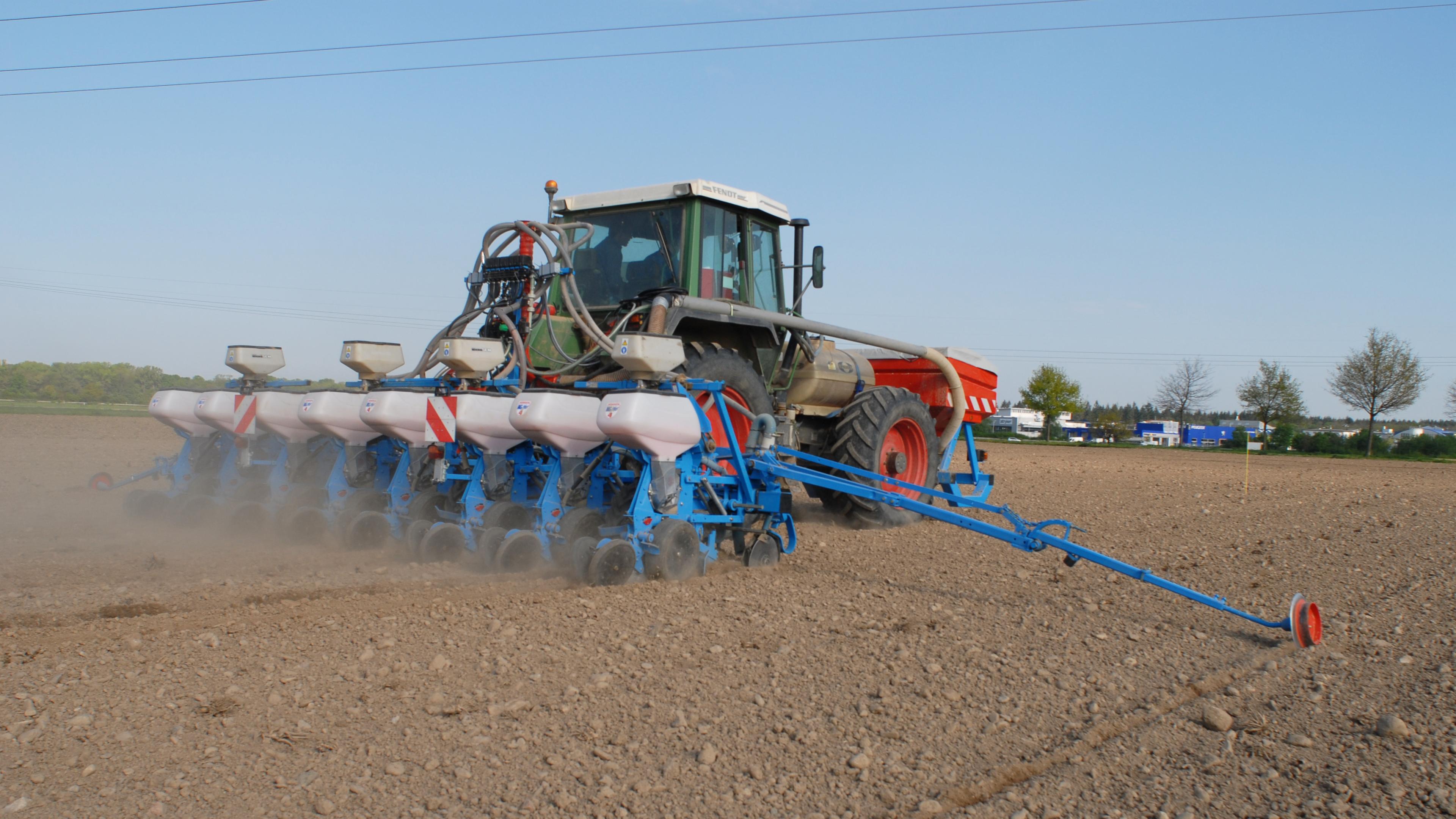 Ein Traktor mit Sämaschine sät auf einem staubigen Feld unter strahlend blauem Himmel, im Hintergrund sind Bäume und Gebäude zu sehen.