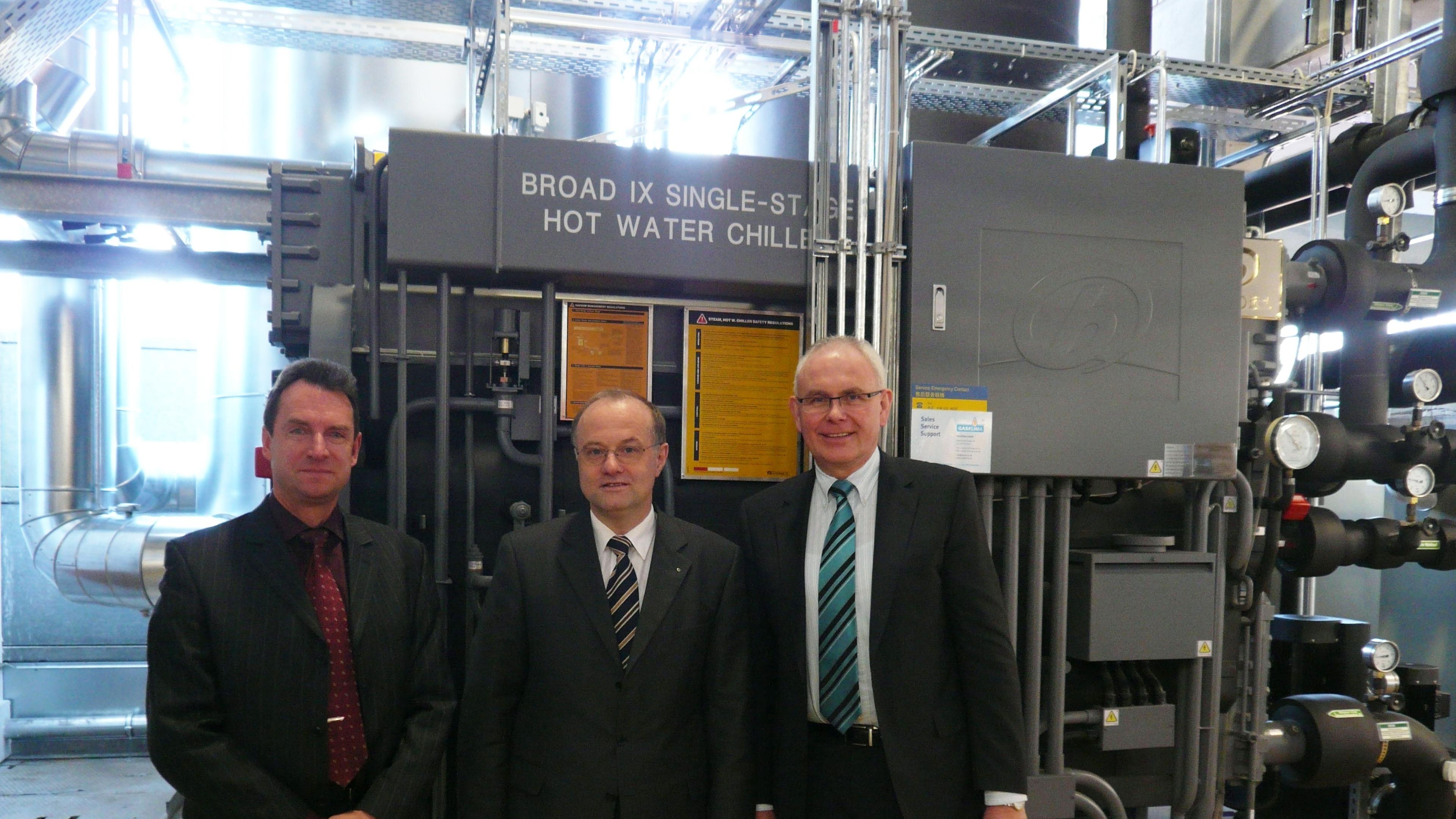 Drei Männer in Anzügen stehen vor einem „Broad IX Single-Stage Hot Water Chiller“ in einer Industrieumgebung.