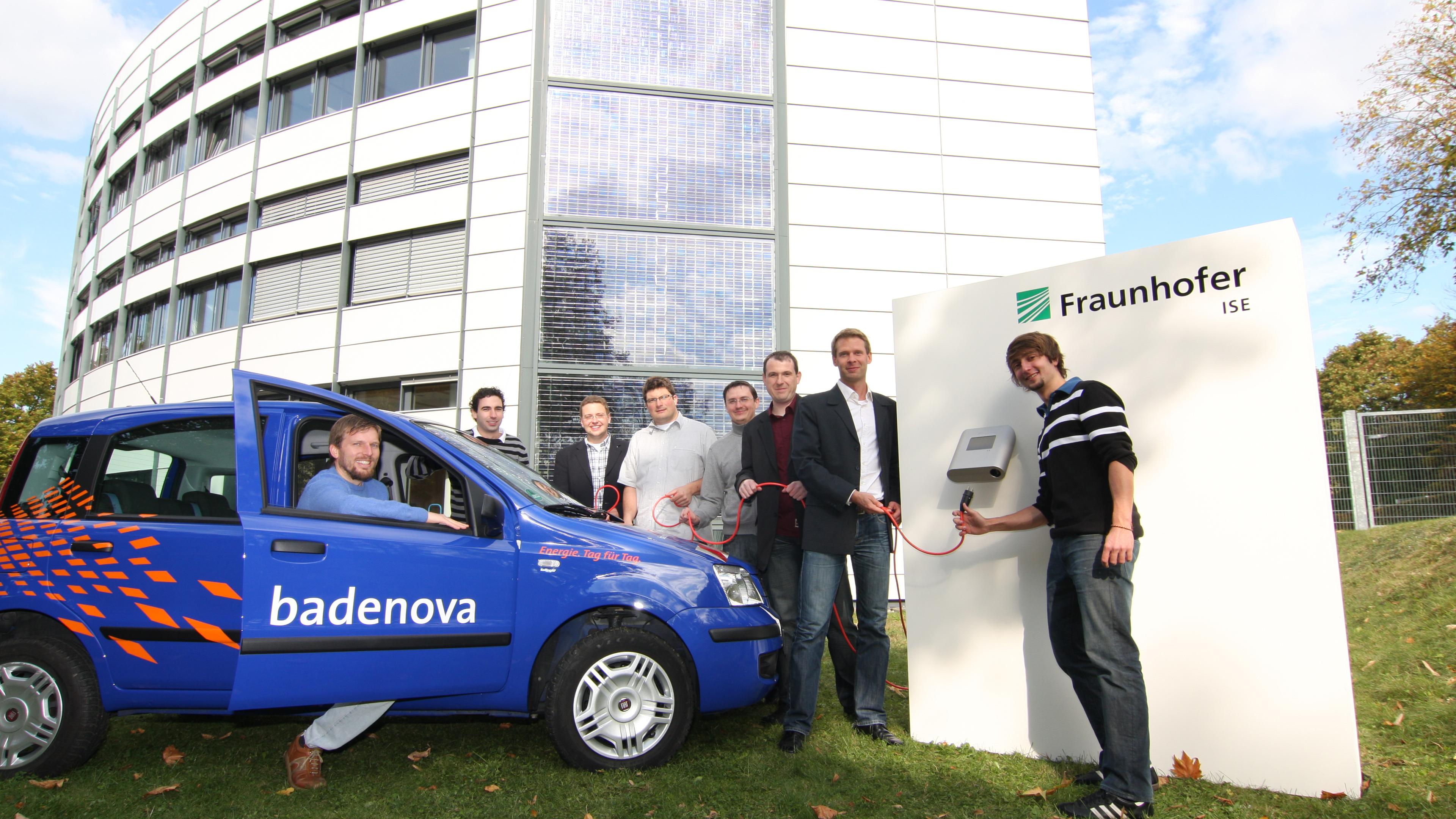 Eine Gruppe von Menschen steht neben einem blauen „Badenova“-Auto und einer Ladestation am Fraunhofer ISE, im Hintergrund ist ein modernes Gebaeude zu sehen.