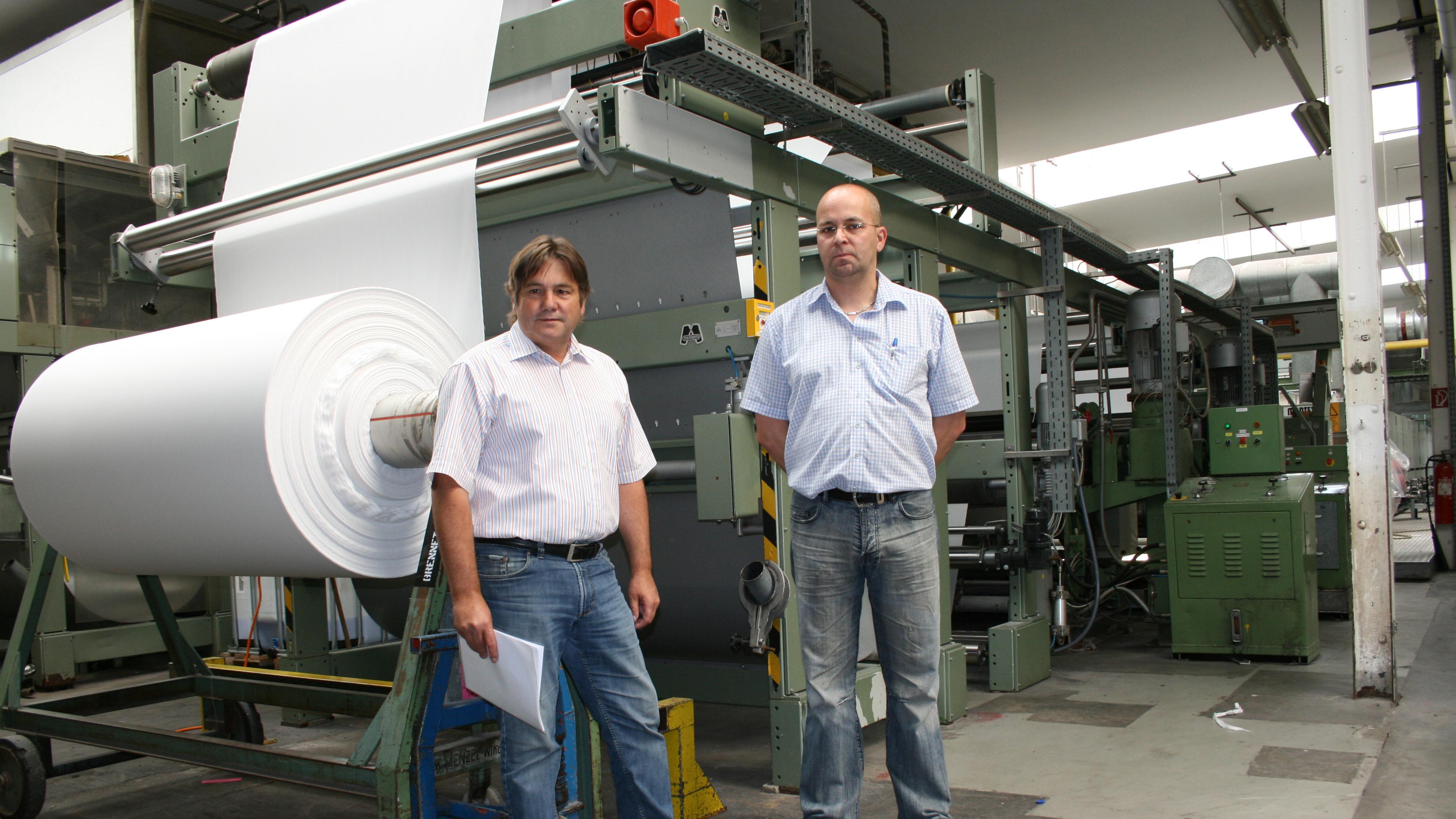 Zwei Männer stehen in einer Produktionshalle vor großen Papierrollen und Industriemaschinen.