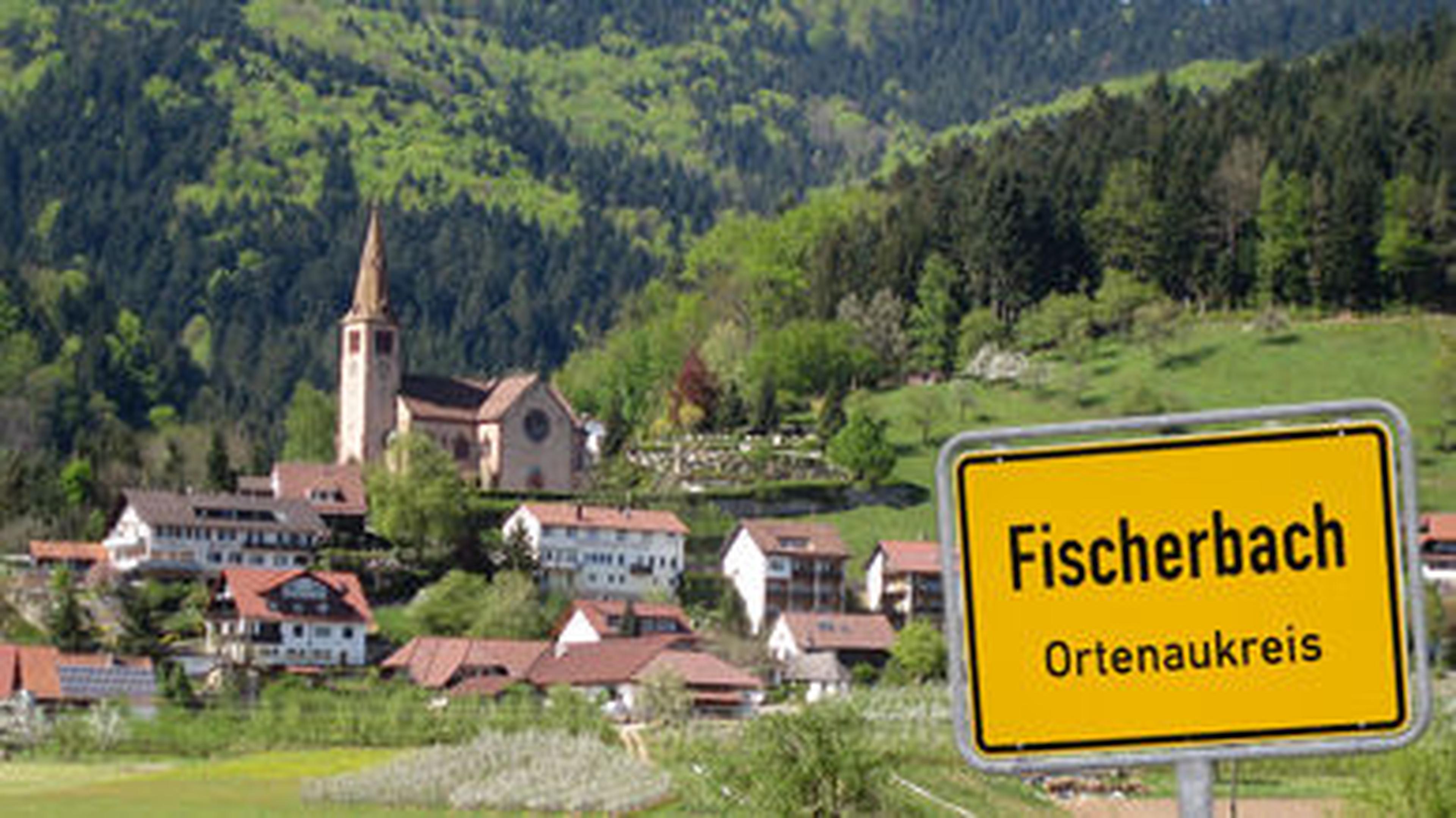Panoramablick auf Fischerbach im Ortenaukreis mit einer Kirche, Häusern und üppig grünen Hügeln im Hintergrund. Im Vordergrund ist ein Ortsschild zu sehen.
