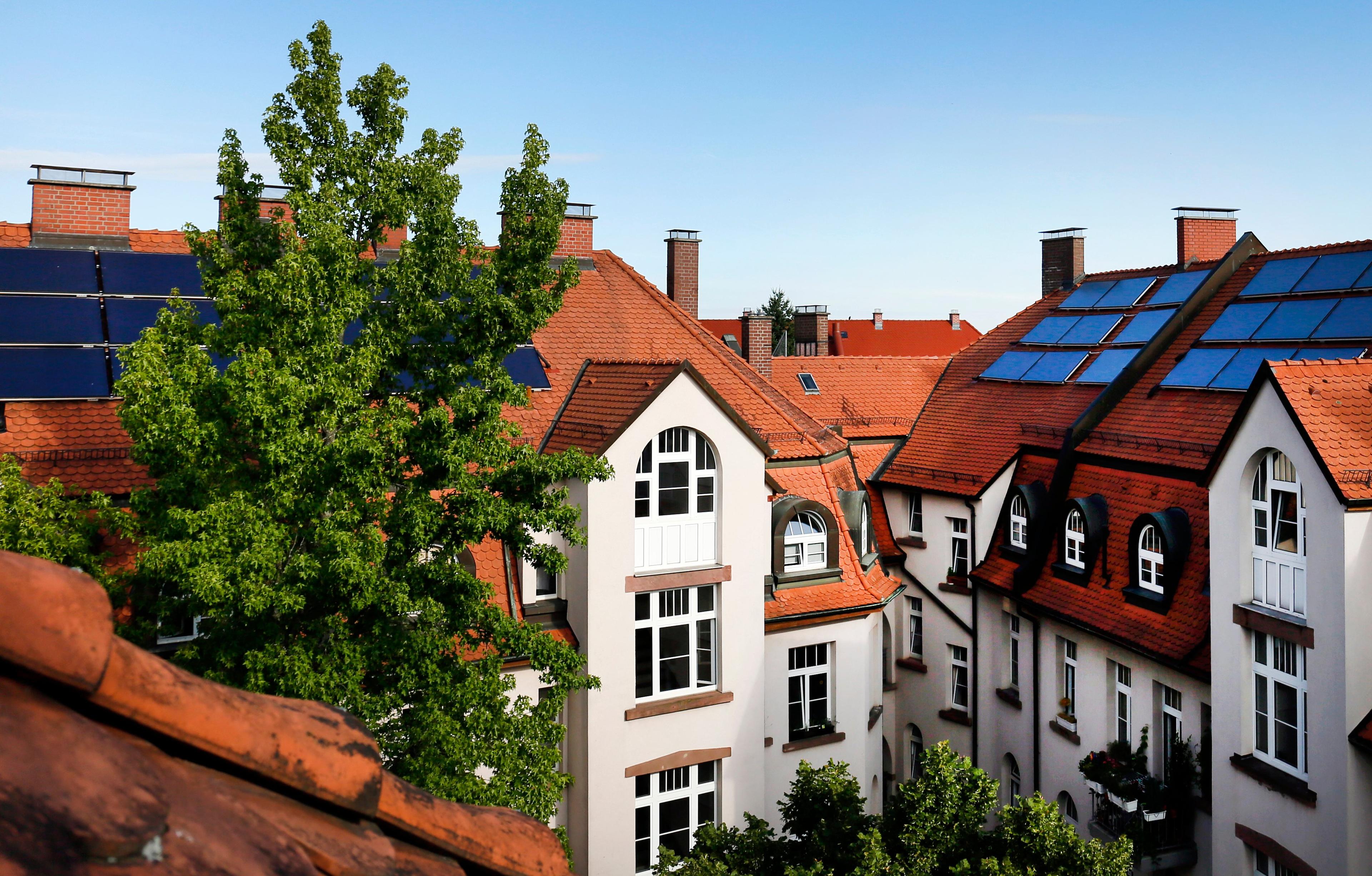 Häuser mit roten Dächern, weißen Wänden und Sonnenkollektoren, die unter einem strahlend blauen Himmel teilweise von grünen Bäumen verdeckt werden.