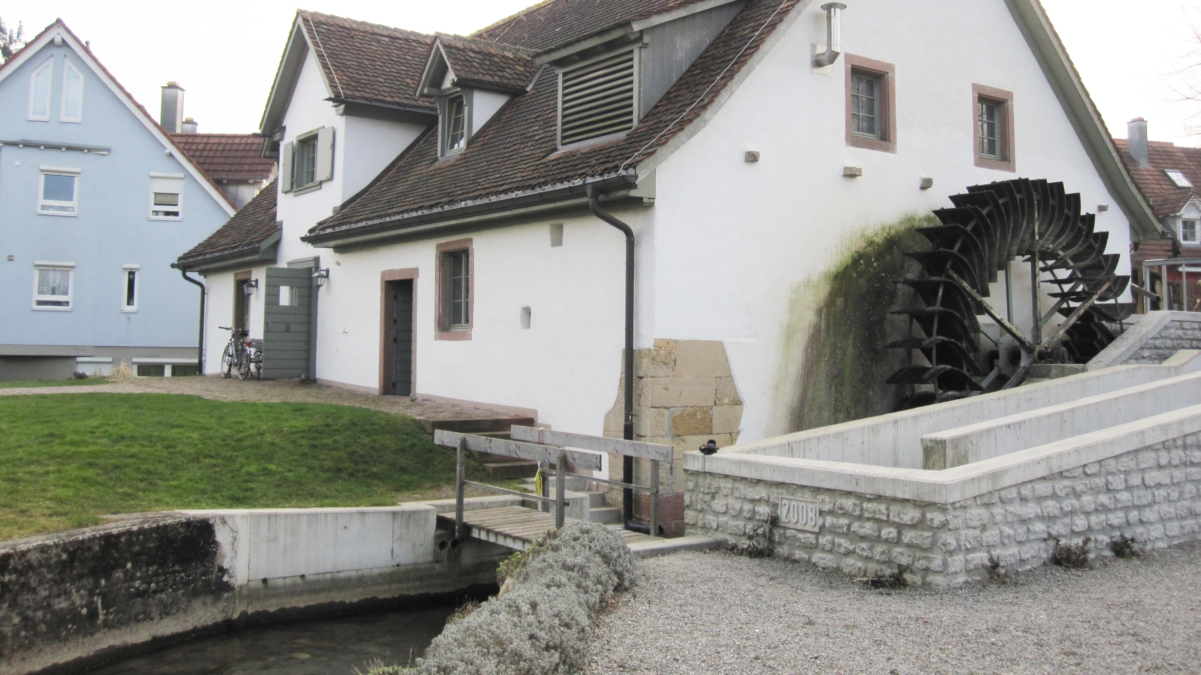 Ein weißes Gebäude mit einem großen Wasserrad an der Seite, das an einem kleinen Wasserkanal steht und an ein blaues Haus grenzt.