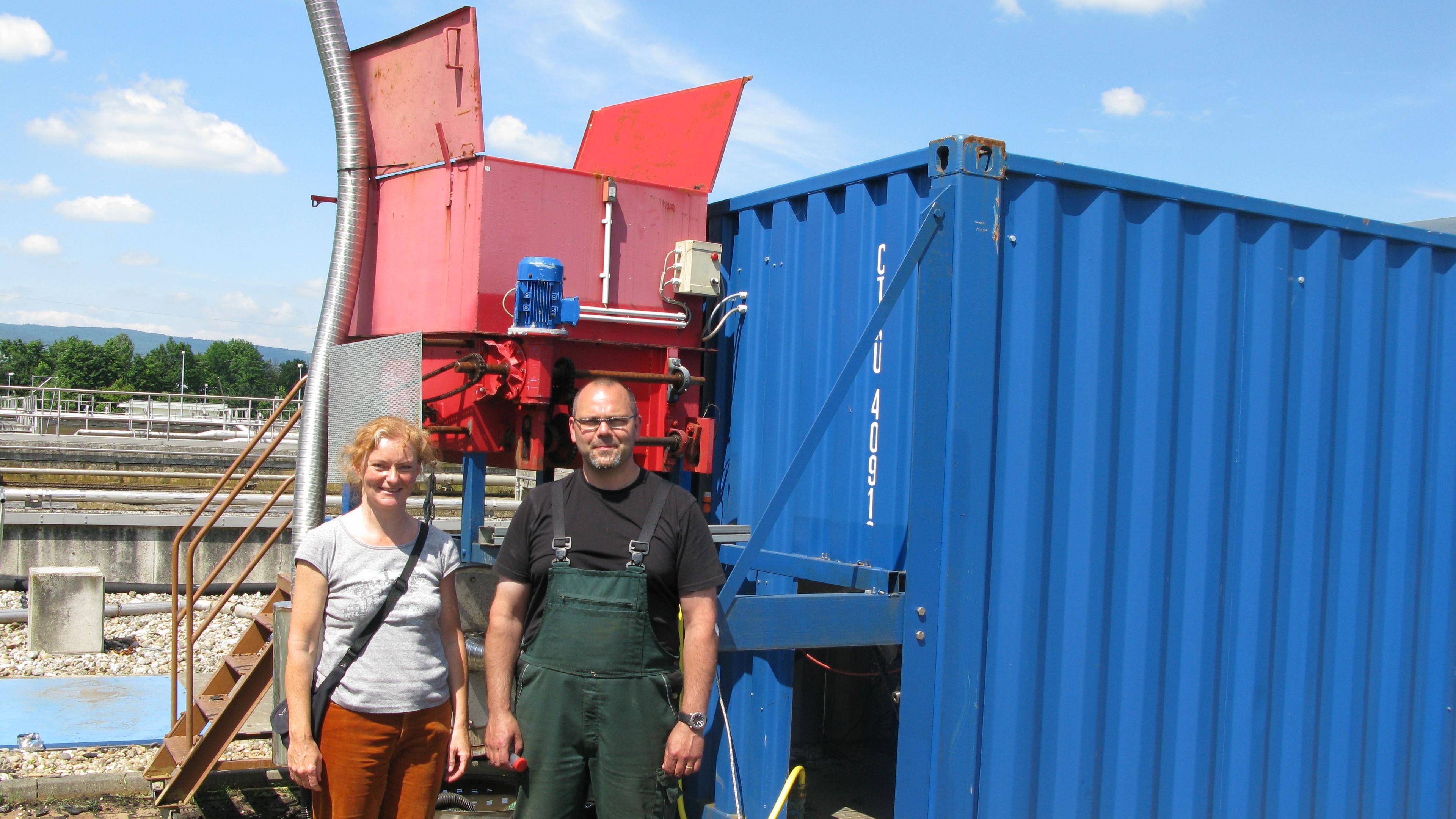Zwei Personen stehen an einem sonnigen Tag im Freien neben einer roten Industriemaschine und einem blauen Container.