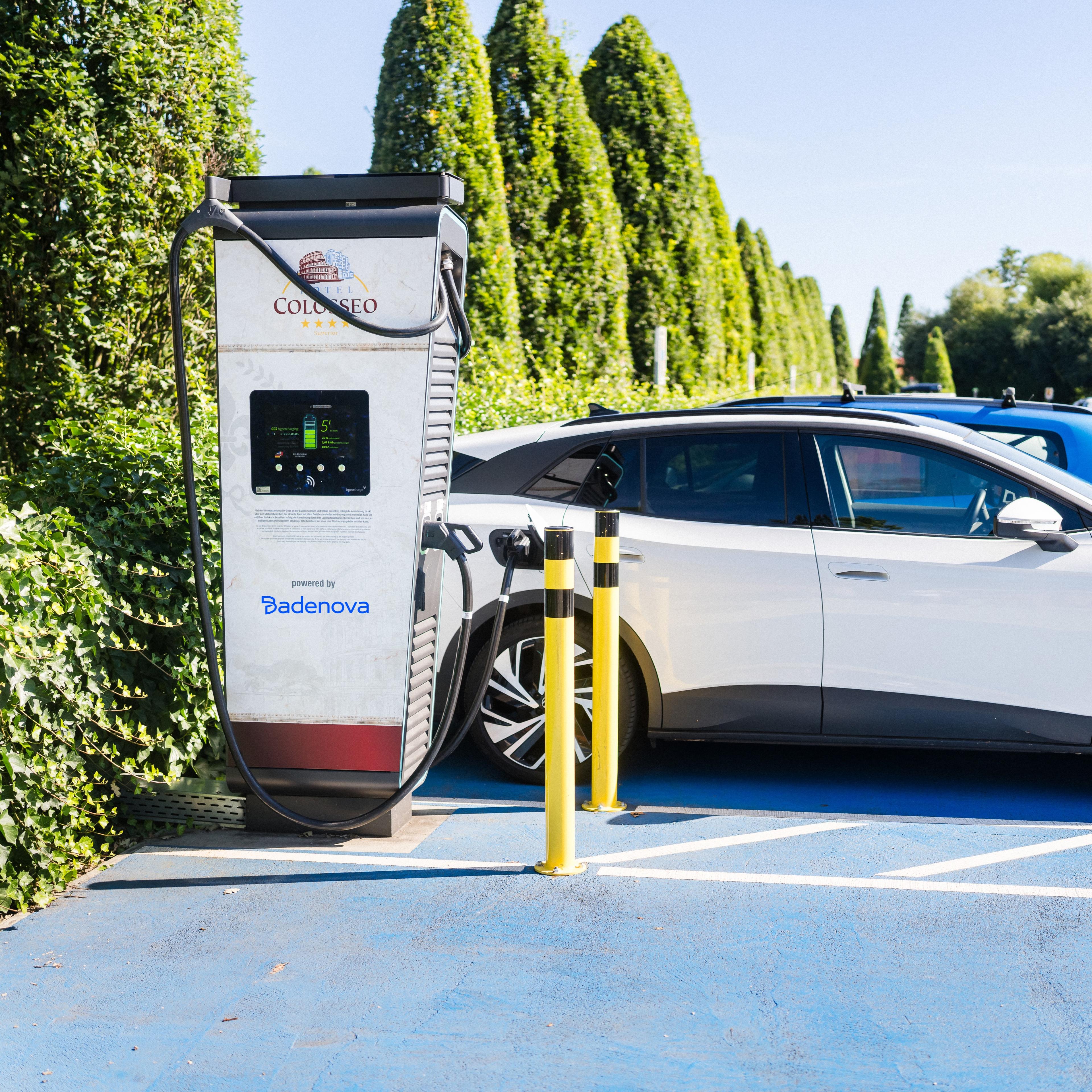 Ein Elektroauto wird an einer Ladestation auf einem Parkplatz aufgeladen, im Hintergrund sind üppige Vegetation und Bäume zu sehen.