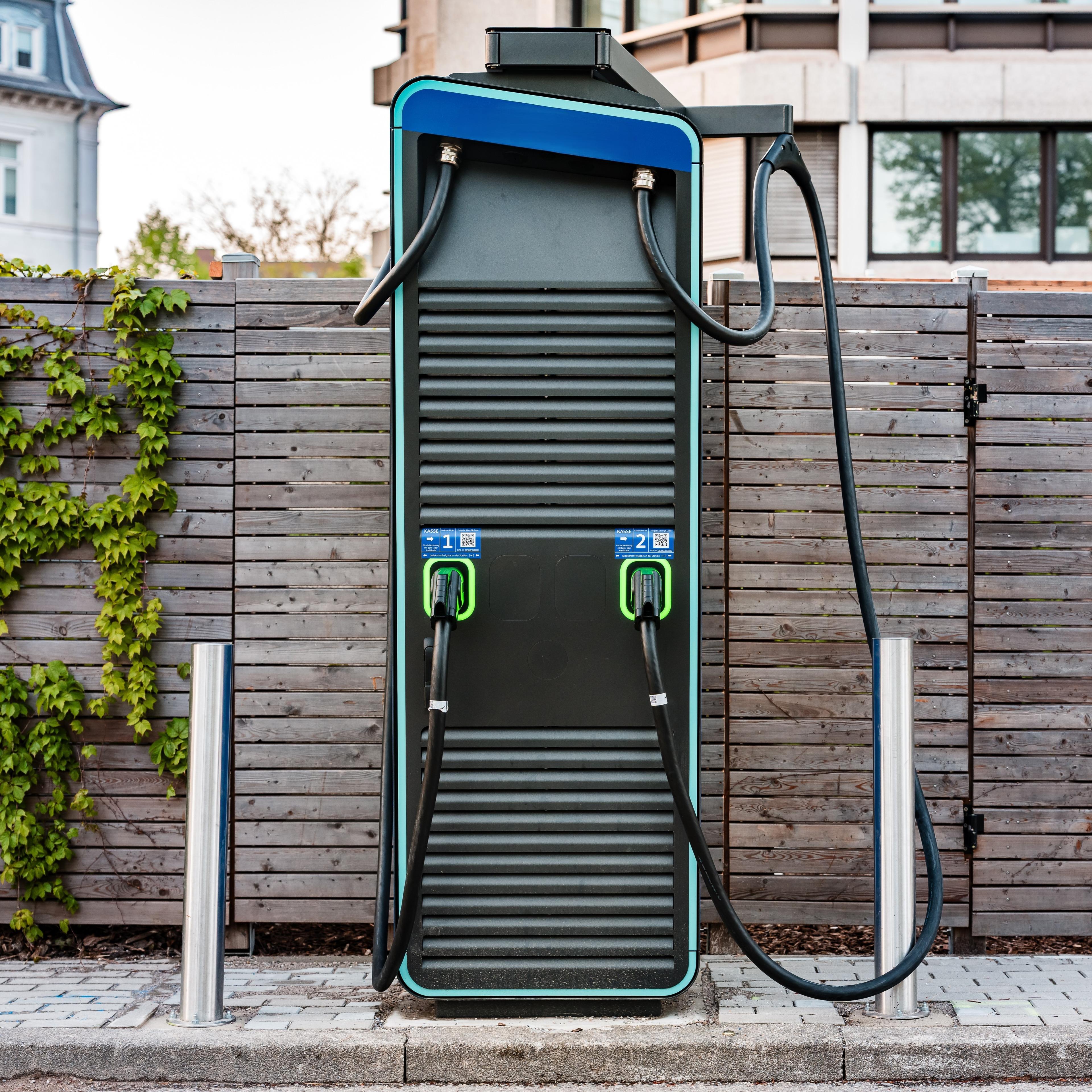 Frontansicht einer Schnellladesäule mit zwei angeschlagenen Ladekabeln, die von zwei silbernen Pollern links und rechts eingerahmt wird.