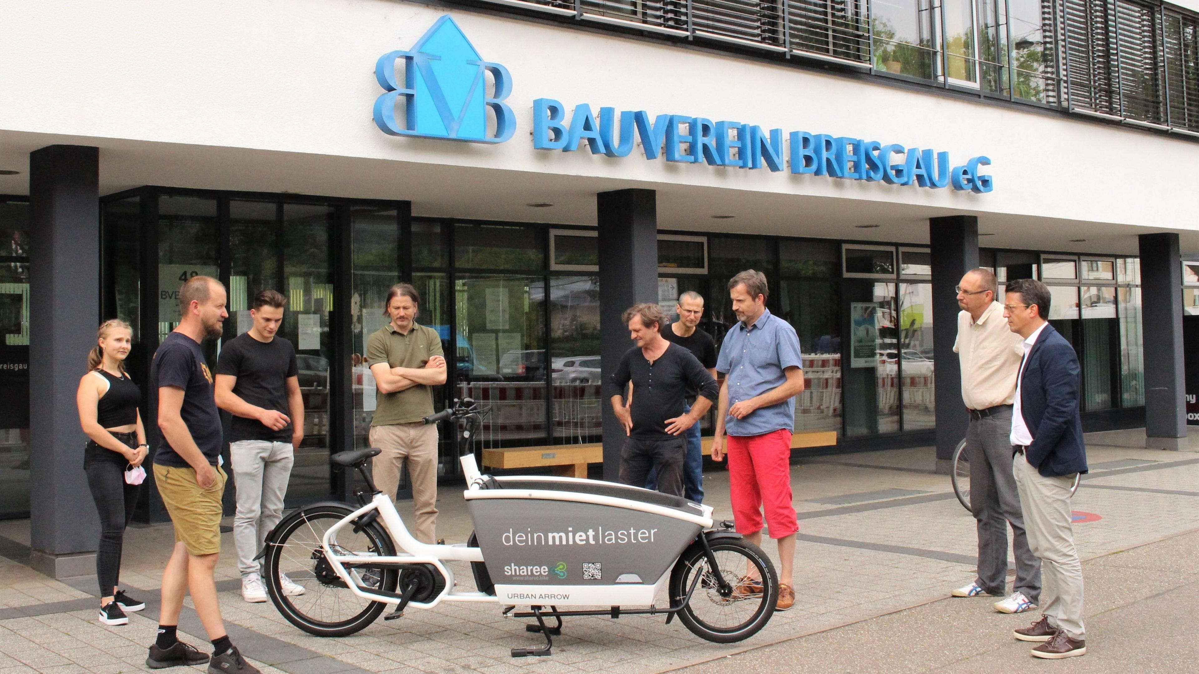 Eine Gruppe von Menschen steht vor einem Gebäude mit der Aufschrift „Bauverein Breisgau eG“ und hat sich um ein elektrisches Lastenfahrrad versammelt.