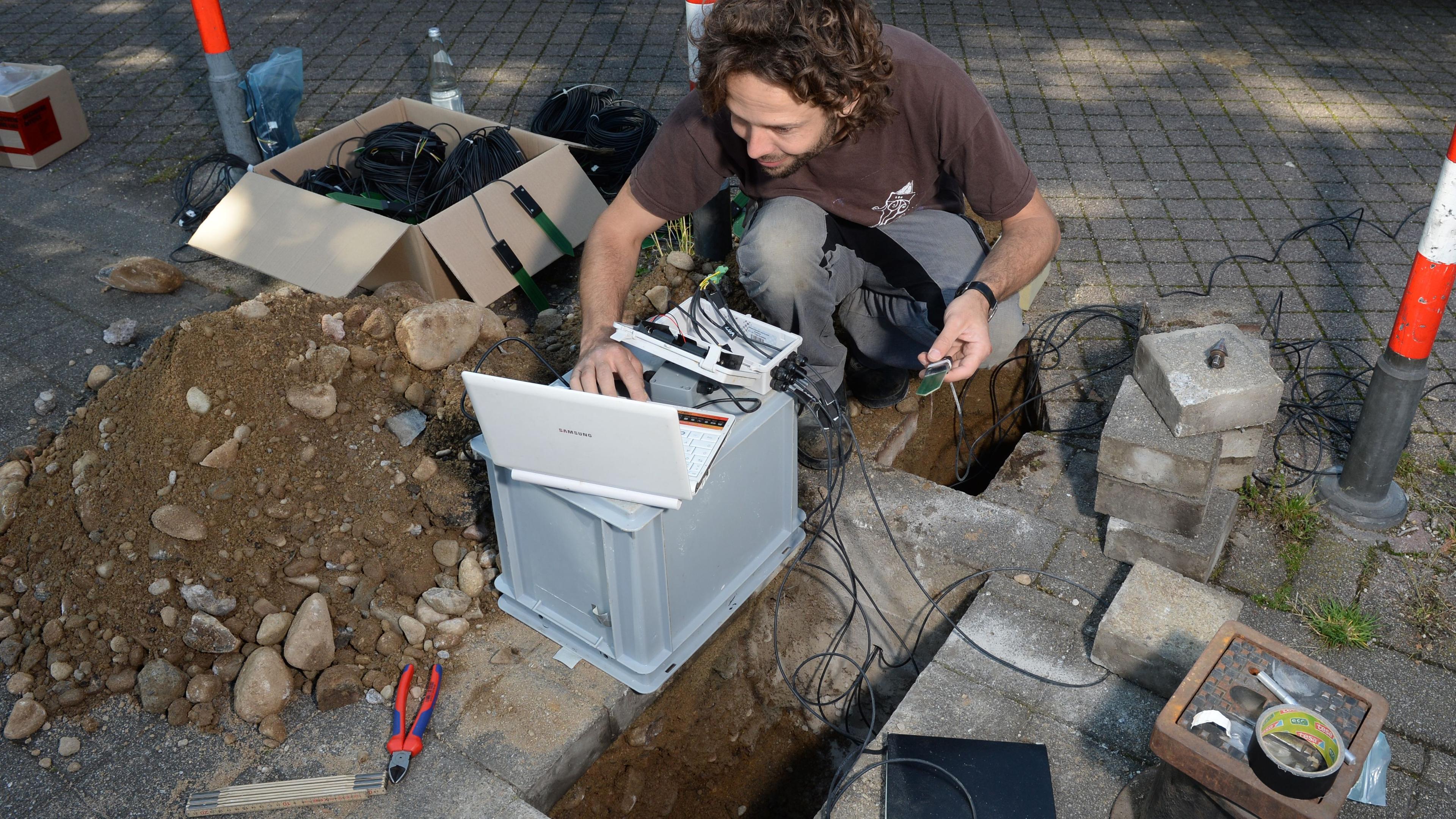 Person, die an einem Laptop arbeitet, der an Kabeln angeschlossen ist, in der Nähe eines offenen Grabens auf einer gepflasterten Fläche, mit herumliegenden Werkzeugen und Geräten.