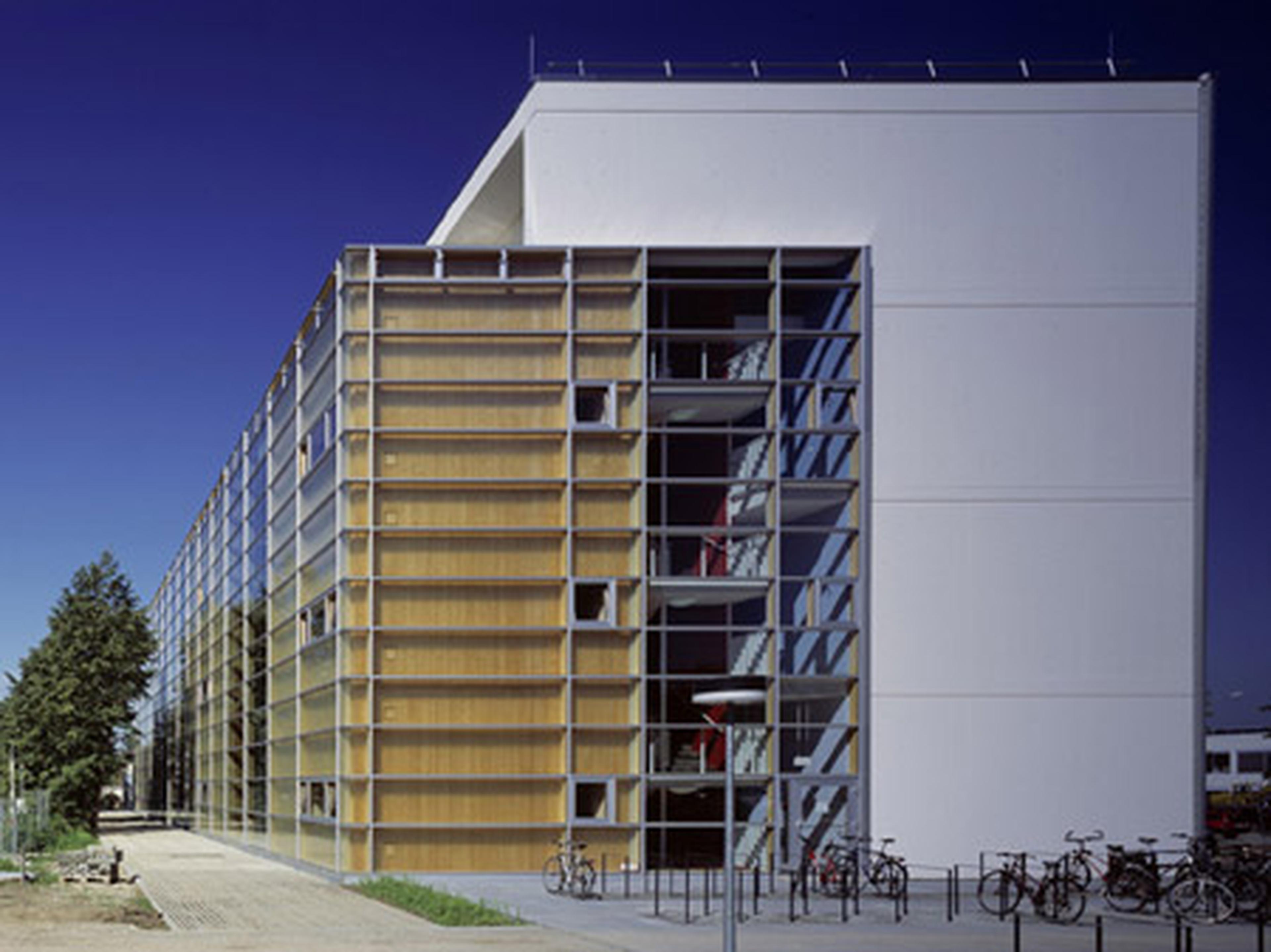 Ein modernes Gebäude mit Holzfassade und Glaselementen, davor stehen Fahrräder, unter strahlend blauem Himmel.