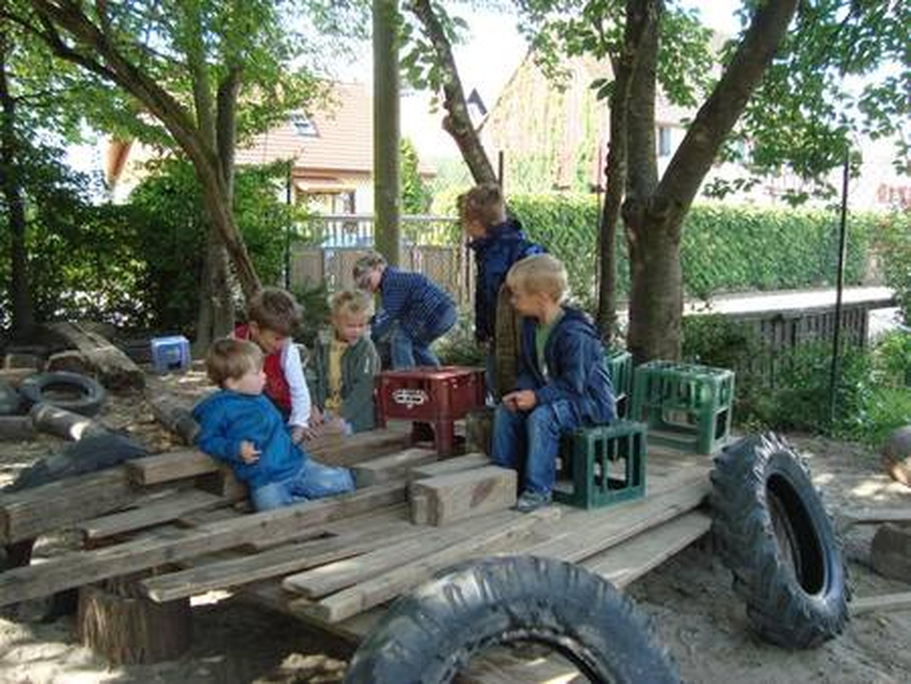 Kinder spielen auf einer provisorischen Holzkonstruktion aus Kisten und Reifen in einer schattigen Außenanlage, umgeben von Bäumen und viel Grün.