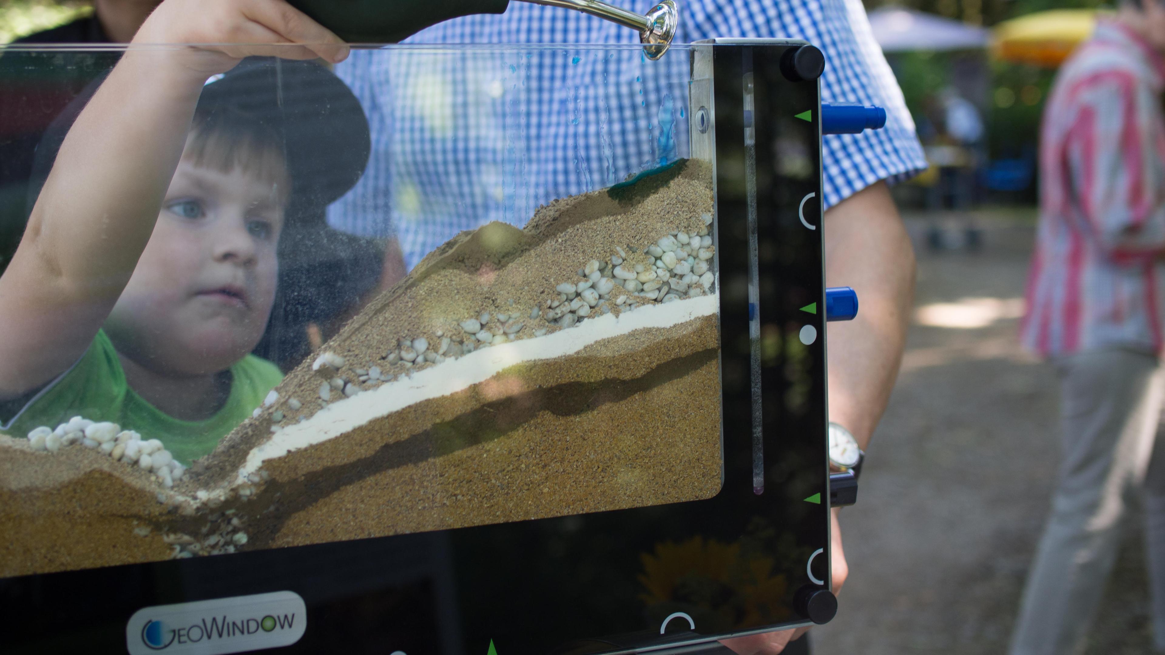 Ein Kind beobachtet ein GeoWindow-Modell, das Schichten aus Sand, Kieselsteinen und Erde zeigt und von einem Erwachsenen im Freien gehalten wird.