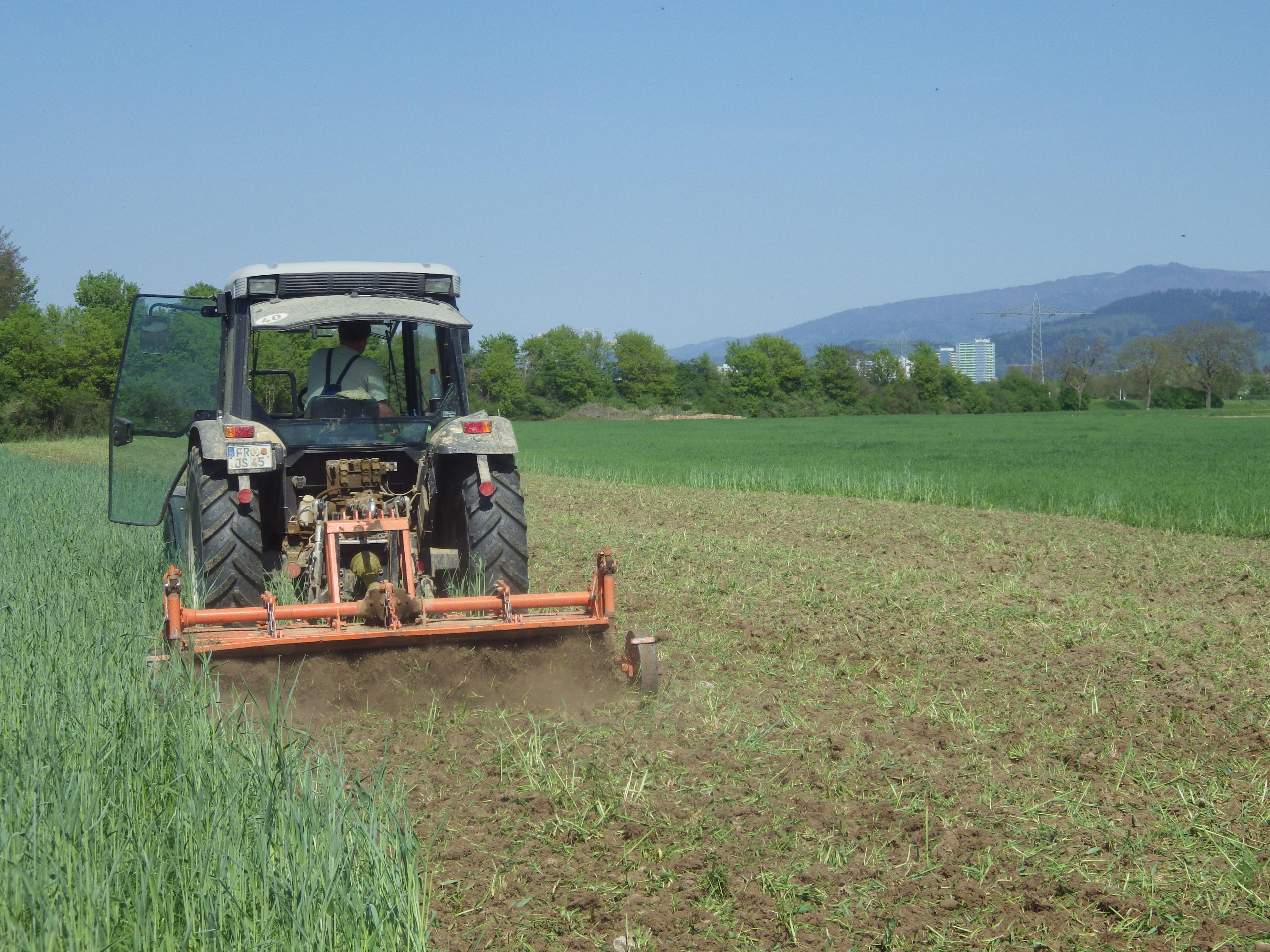 Ein Traktor pflügt ein Feld mit einer grünen Landschaft und fernen Hügeln unter einem klaren blauen Himmel.