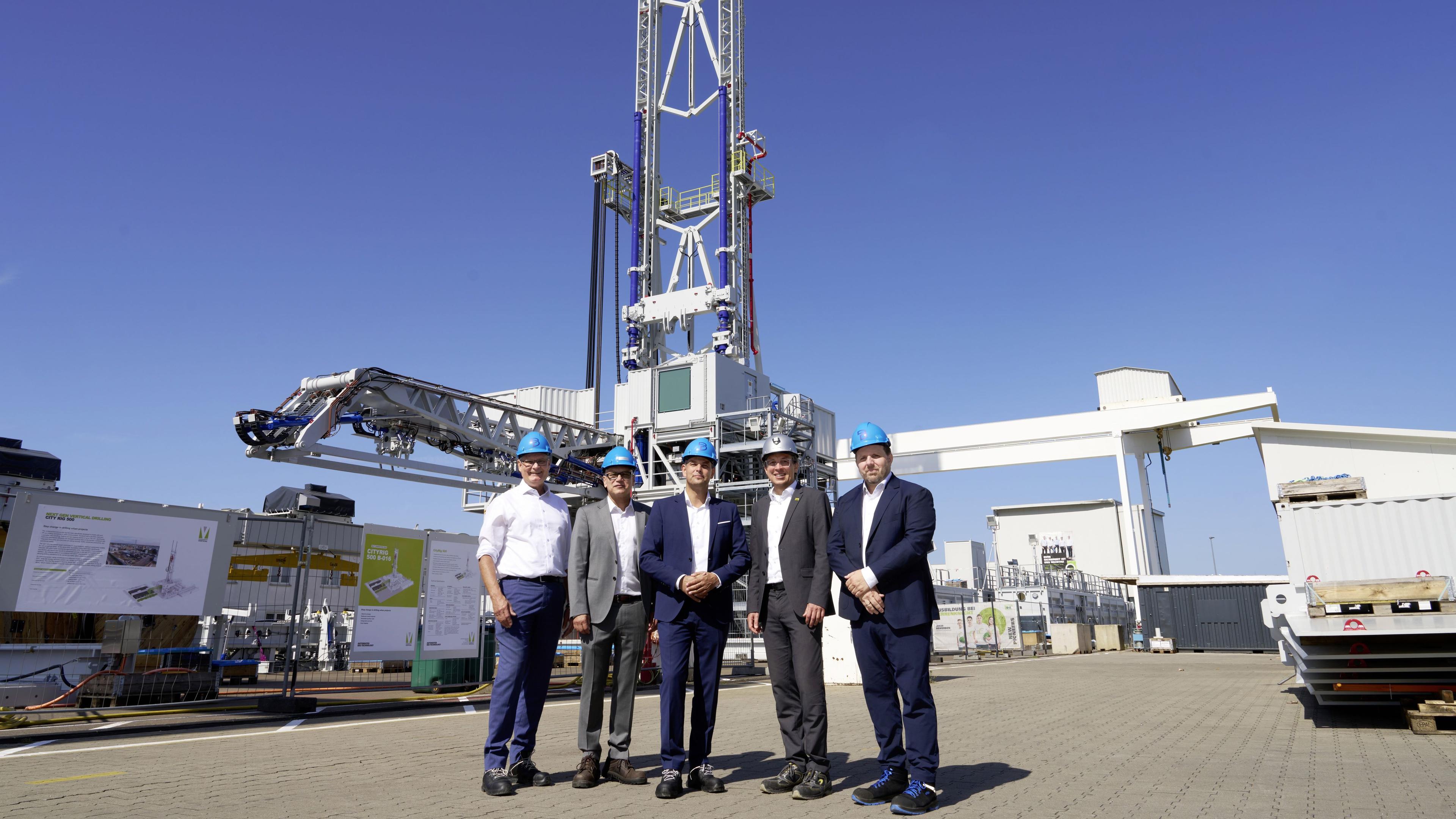 Five men in suits and hard hats stand in front of a tall industrial structure under a clear blue sky.